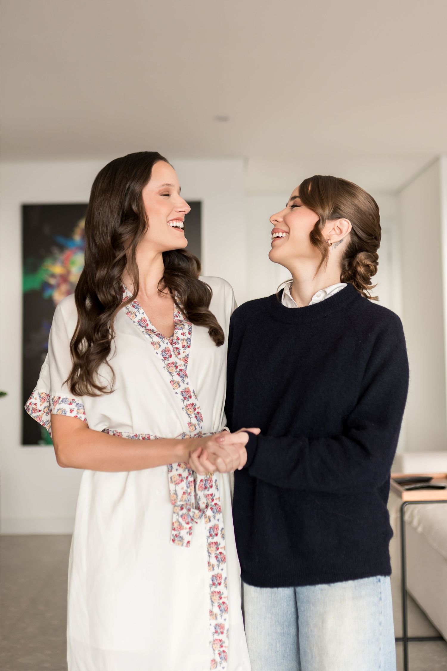 Two smiling women holding hands indoors, one in a floral white dress, the other in a navy sweater and jeans.