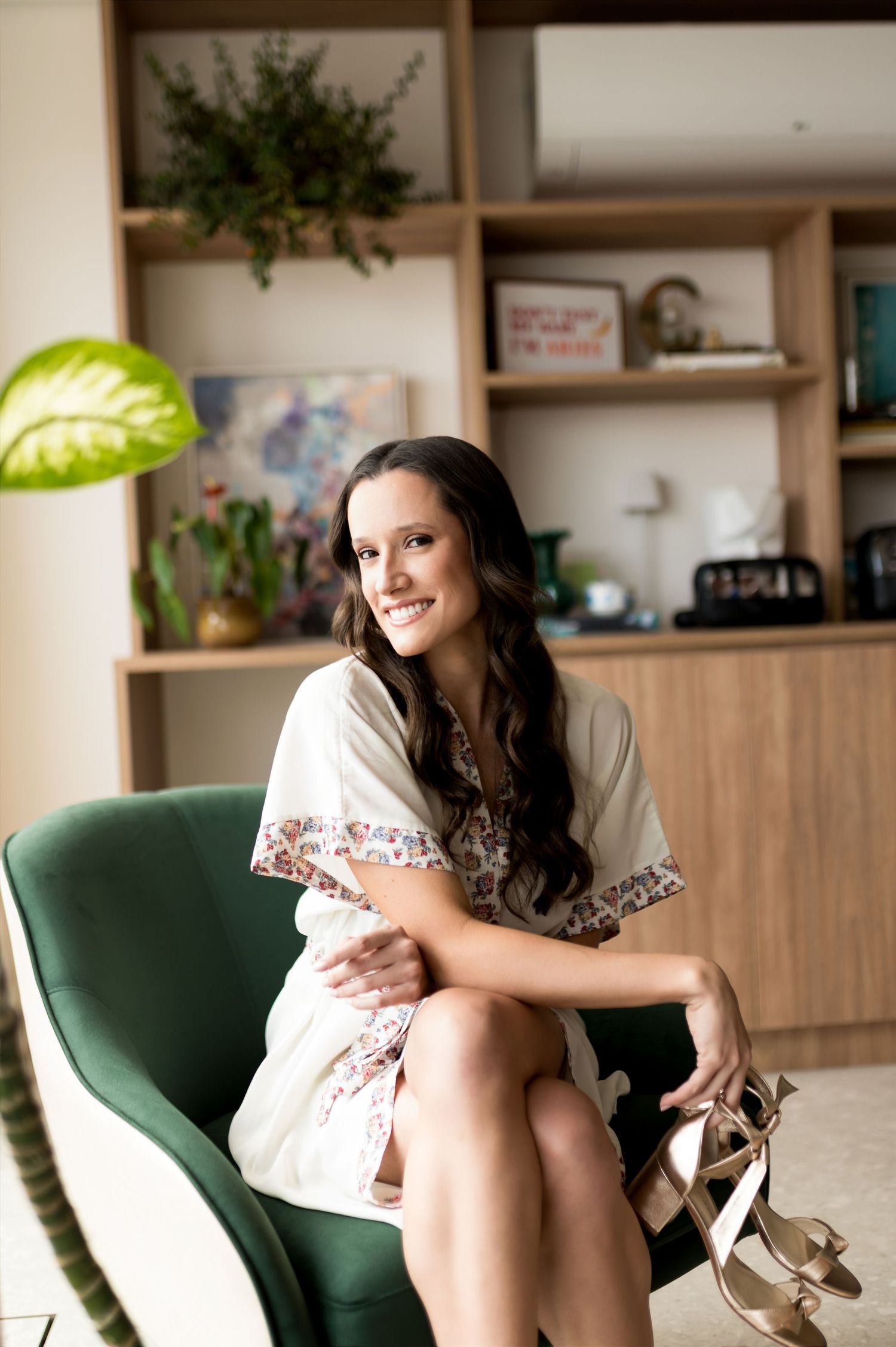 Smiling woman in floral robe sitting in green velvet chair holding heels, with bookshelf and plants in background.