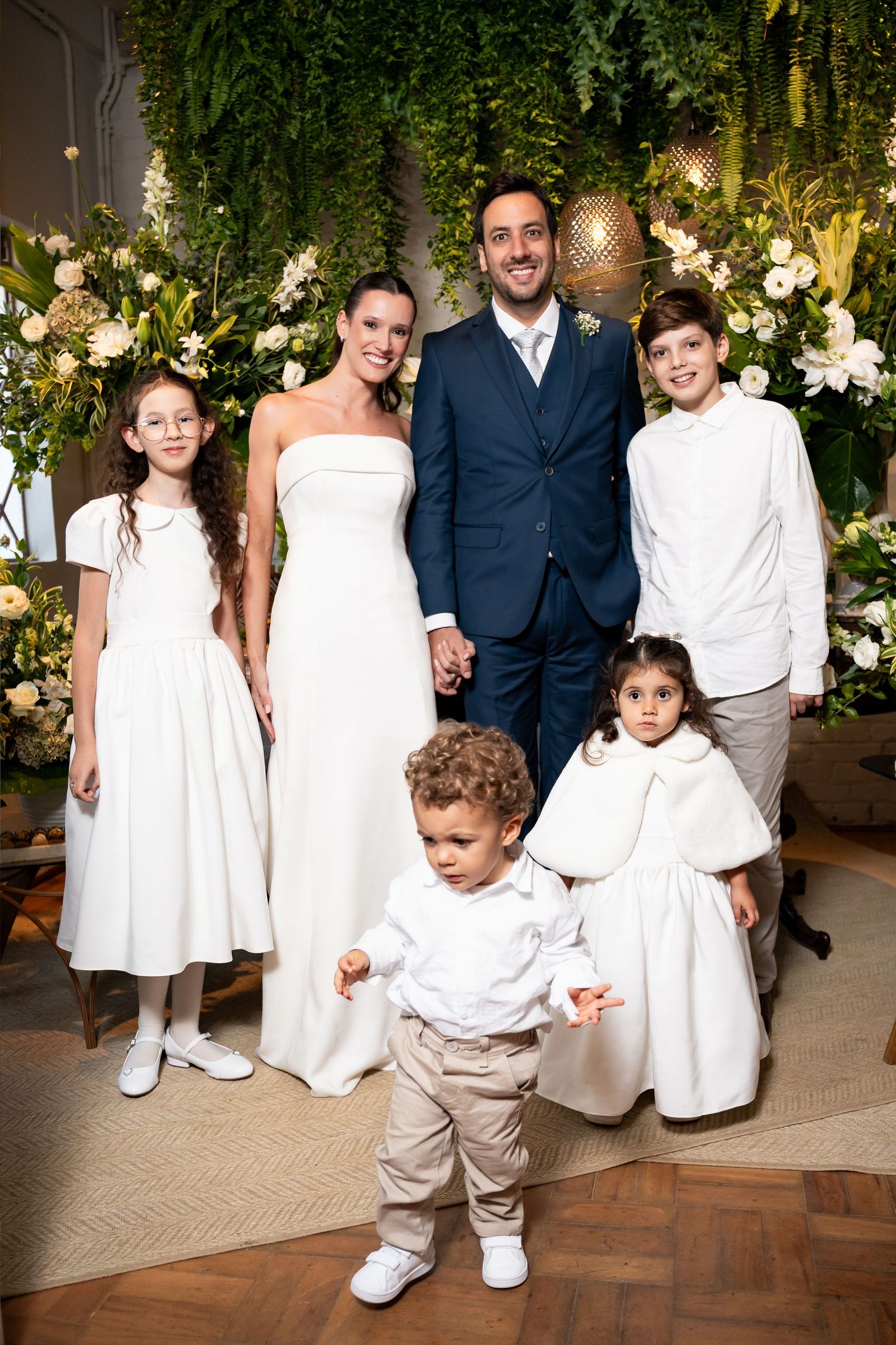 Bride and groom pose with four children in front of lush floral greenery backdrop at elegant wedding ceremony.