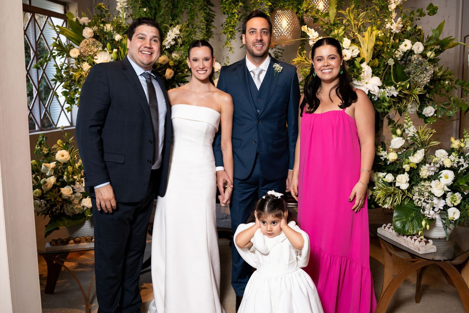 Wedding party poses with bride in white gown, groom in navy suit, flower girl, and guests beside floral arrangements.