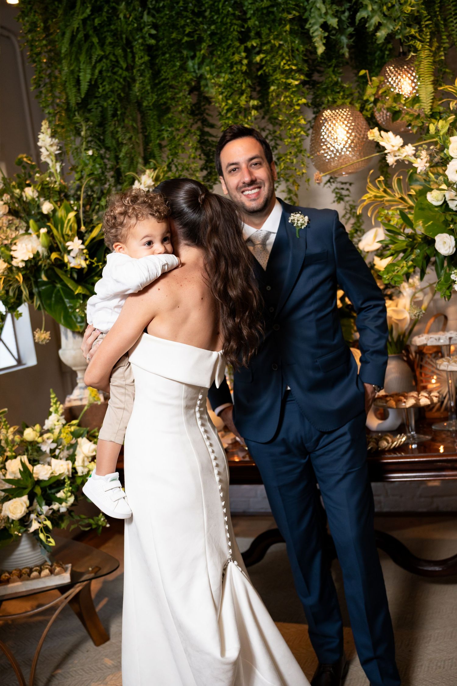 Bride in white gown holds child while groom in navy suit smiles at wedding reception with floral greenery backdrop.