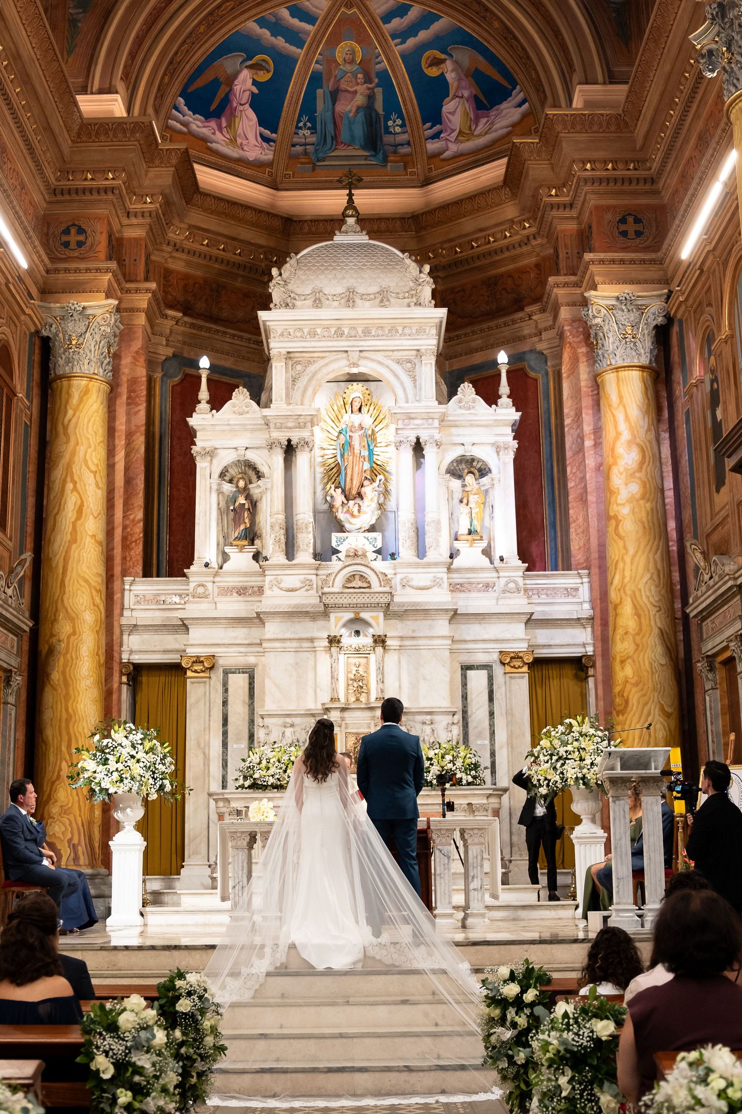 Bride and groom stand before ornate white marble altar in elegant church during wedding ceremony.