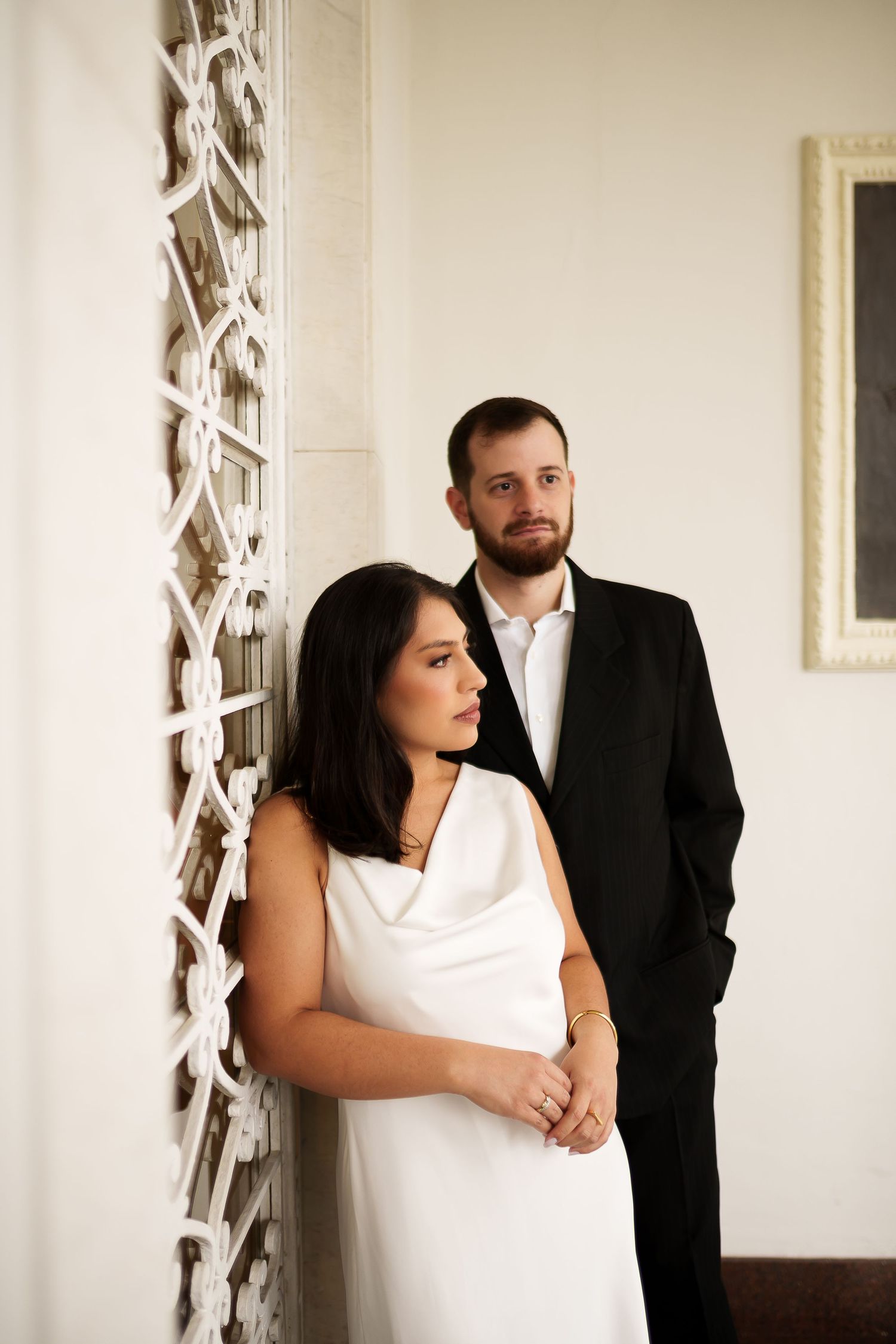 Elegant couple posing indoors, woman in white dress and man in black suit beside decorative white wall.