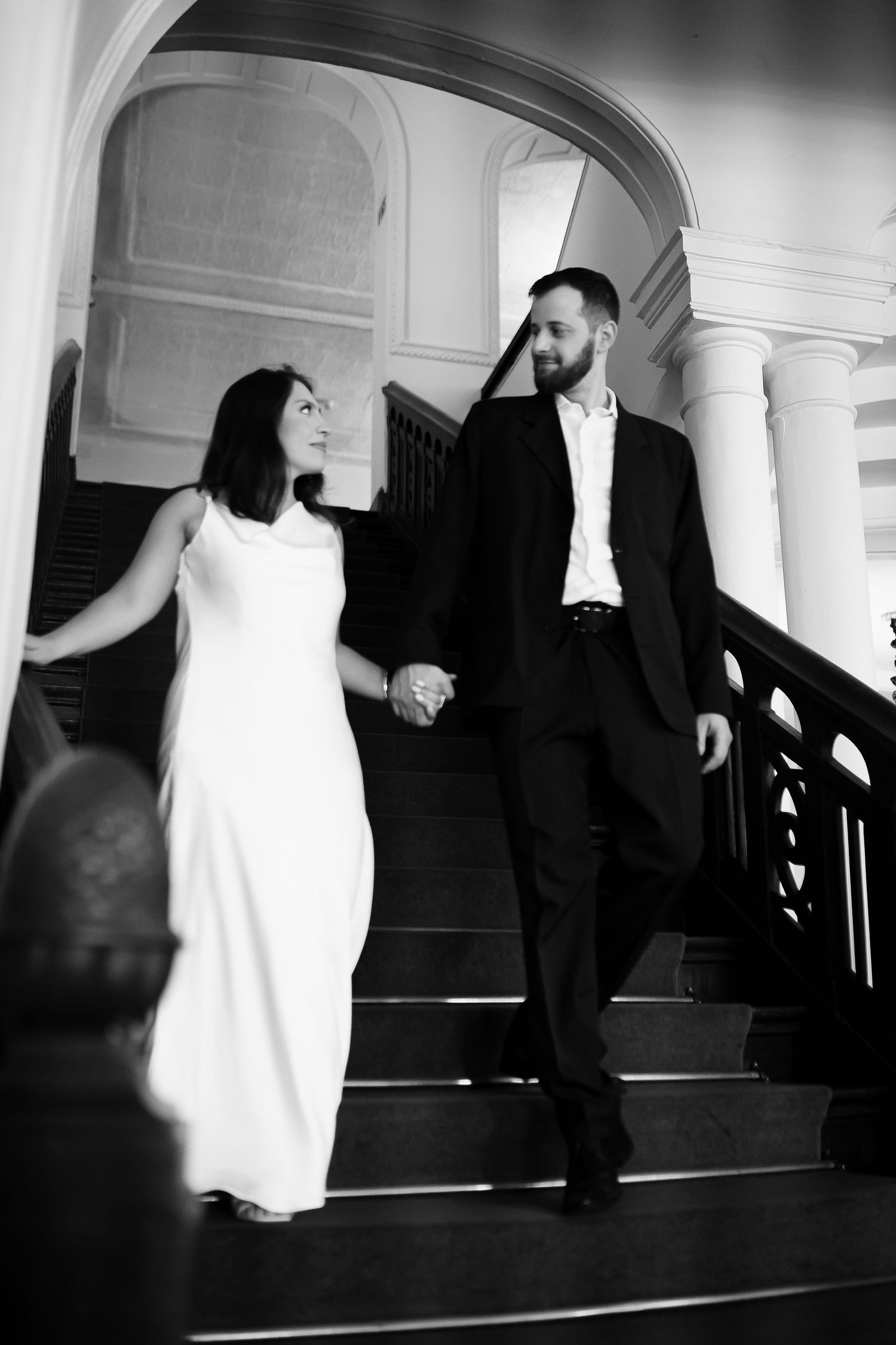 Black and white photo of elegant couple holding hands walking down grand staircase, woman in white gown, man in black suit.