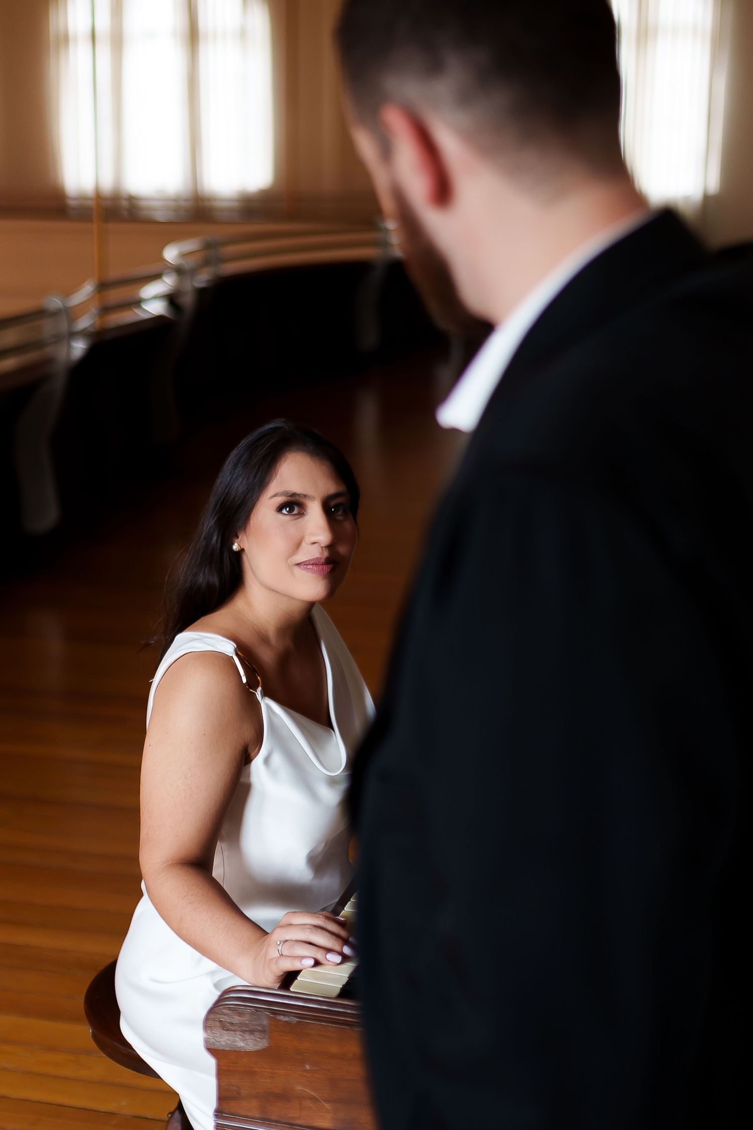 A woman in a white satin dress sits at a piano, gazing up at a man in a black suit in an elegant room.