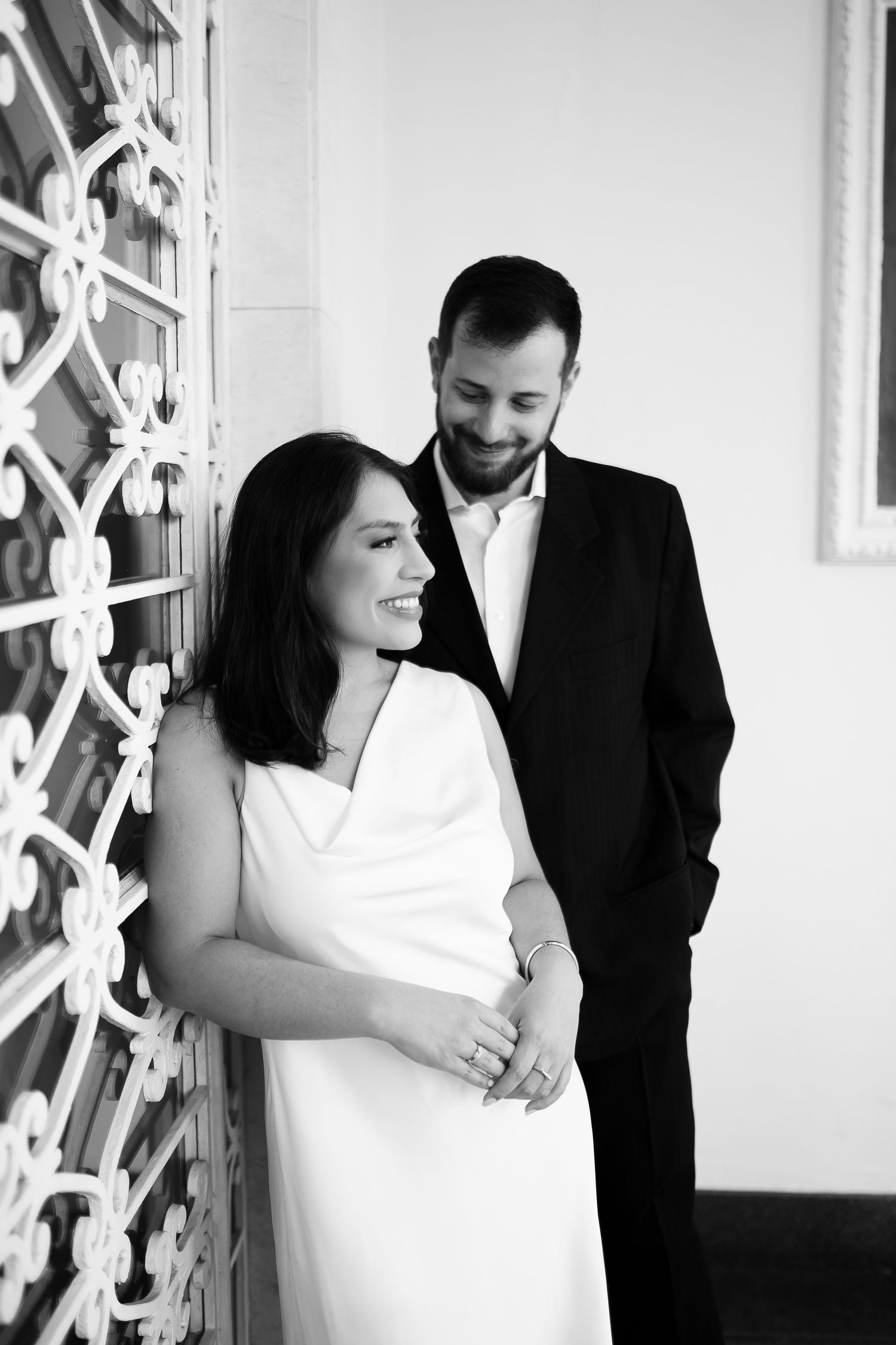 Black and white photo of a smiling bride in white dress and groom in dark suit posing together near ornate iron gate.