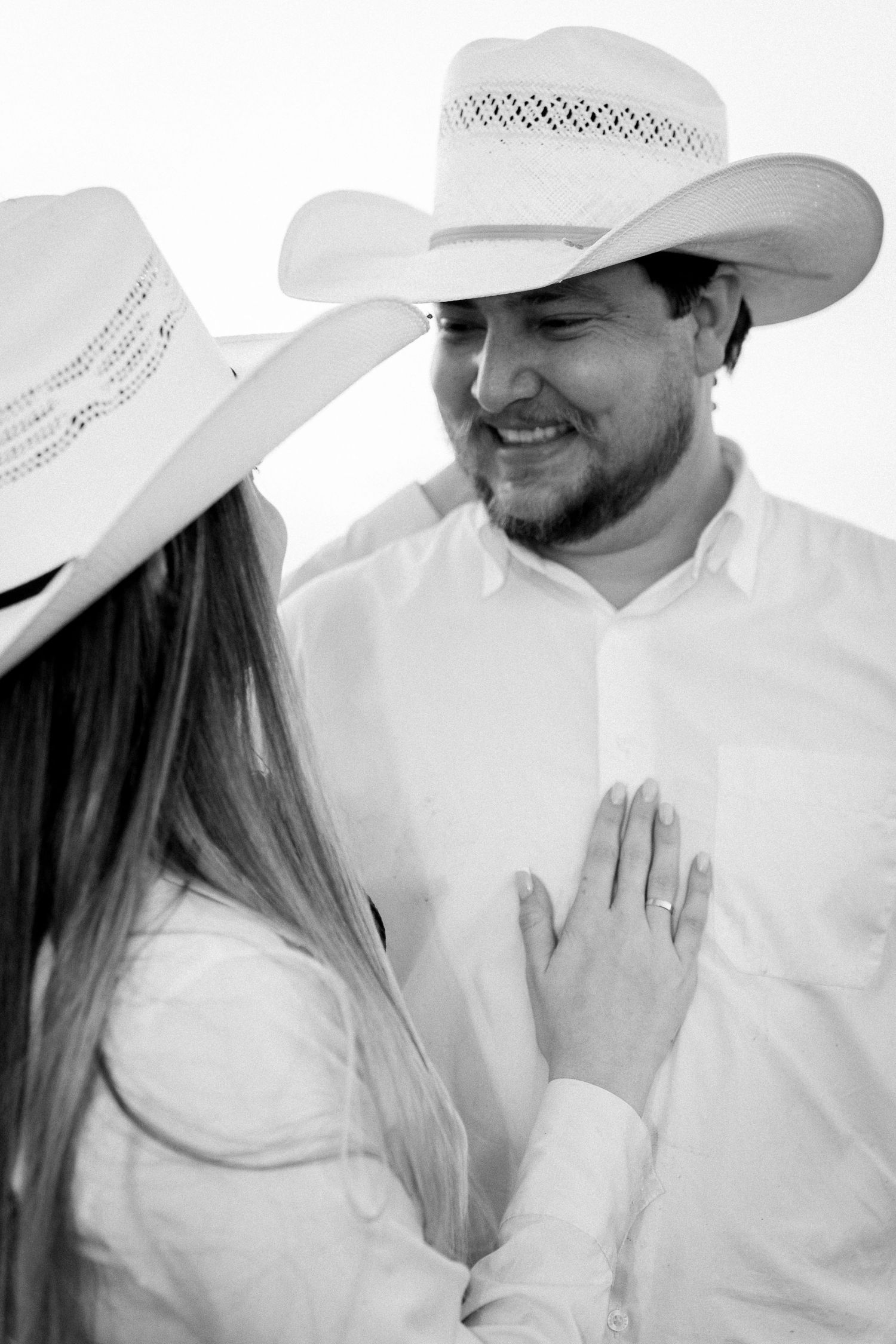 Black and white photo of a couple in white cowboy hats, woman placing hand on mans chest, showing engagement ring.