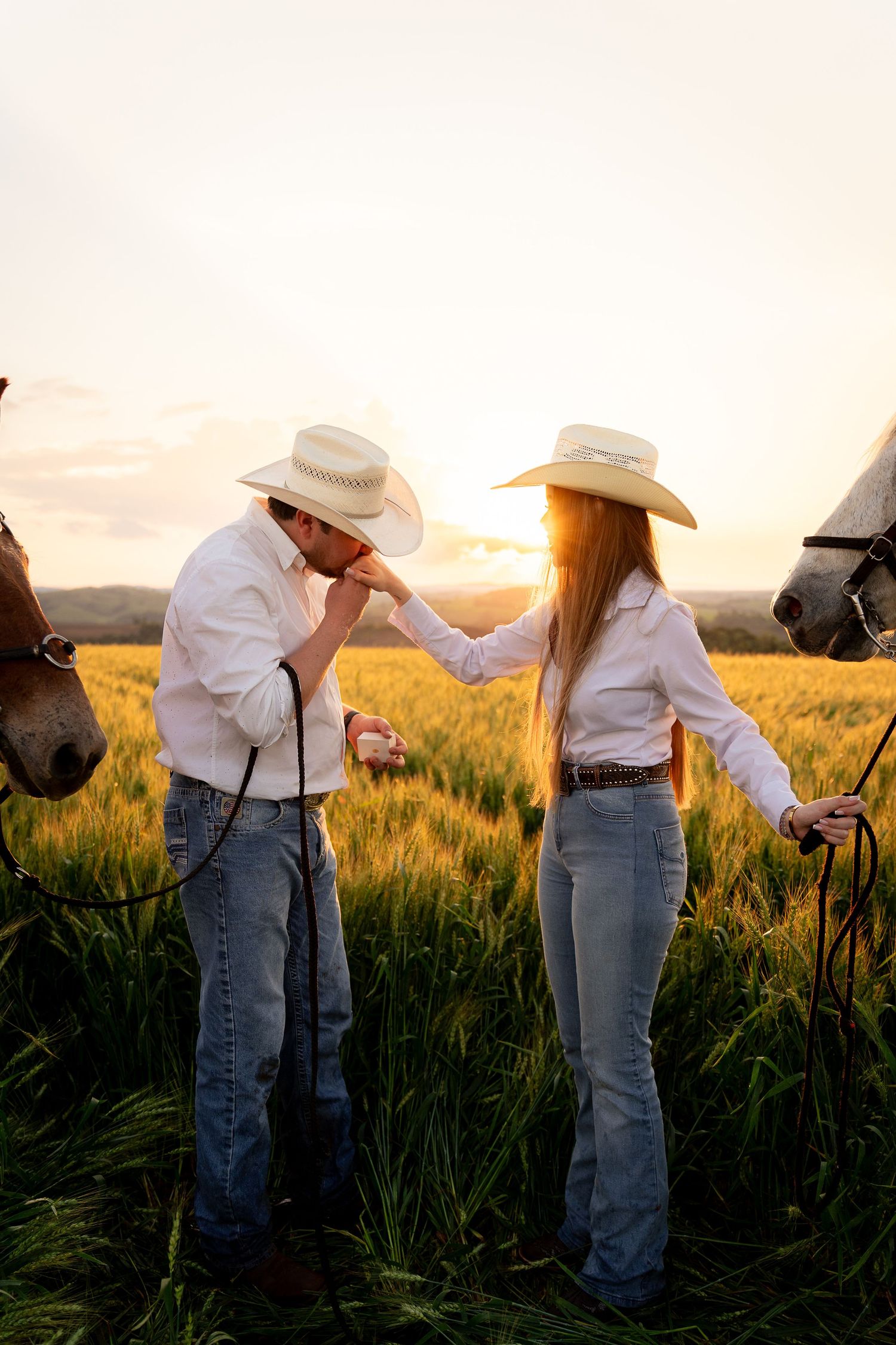 A couple in cowboy hats and jeans share a romantic hand-kiss moment in a golden wheat field at sunset with horses nearby.