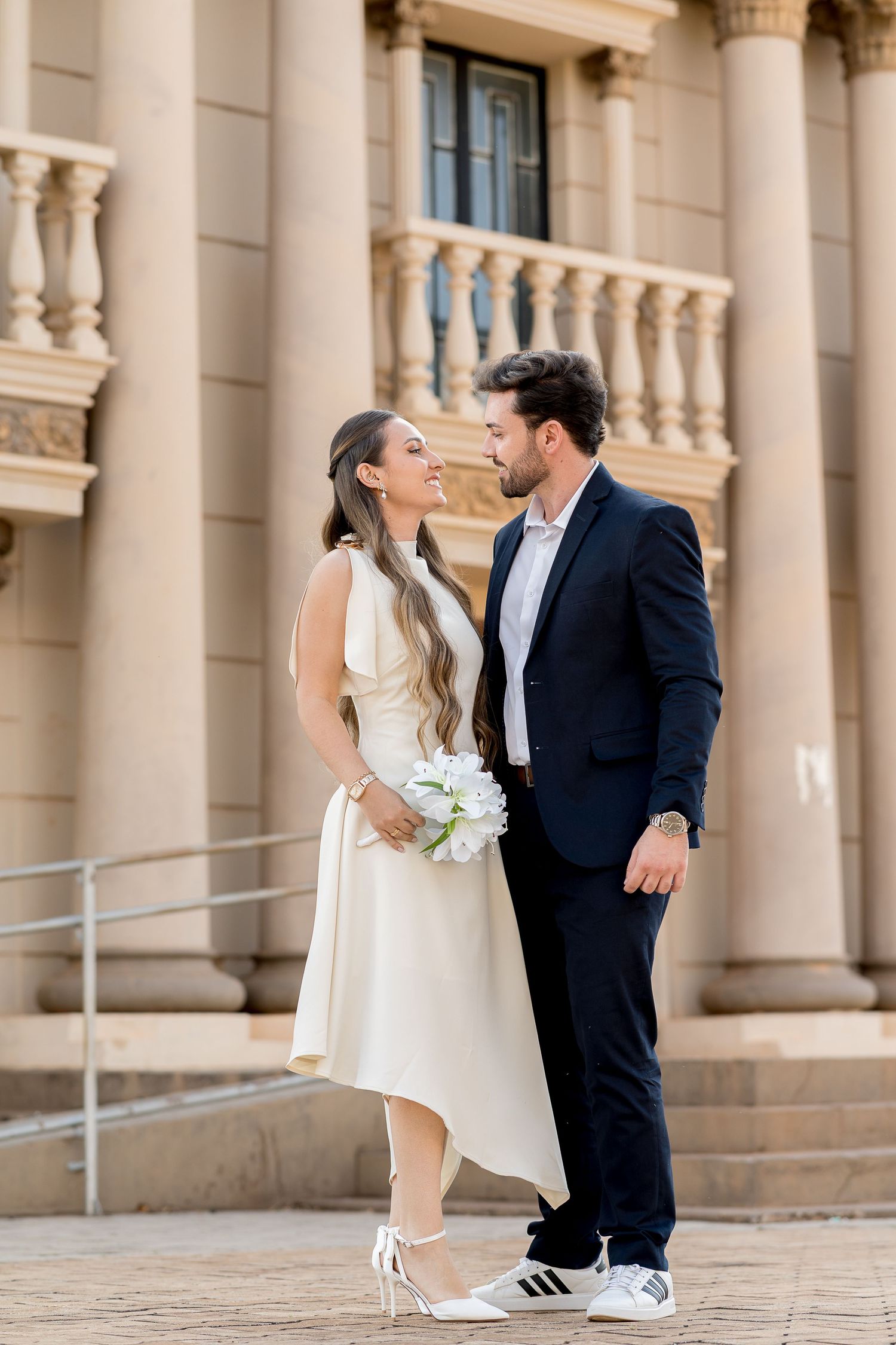 Couple in wedding attire smiling at each other in front of a grand building, bride holding white flowers.