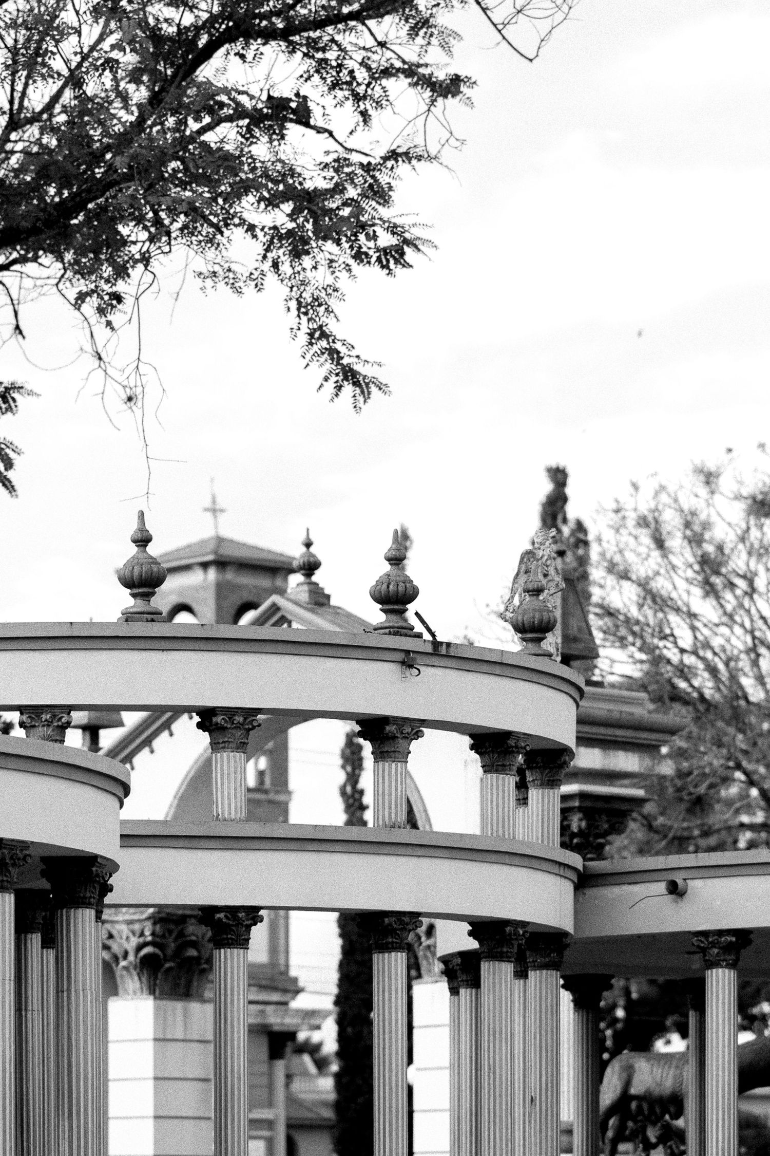 Black and white photo of ornate classical columns and arches at a historic cemetery with statues and cross visible.