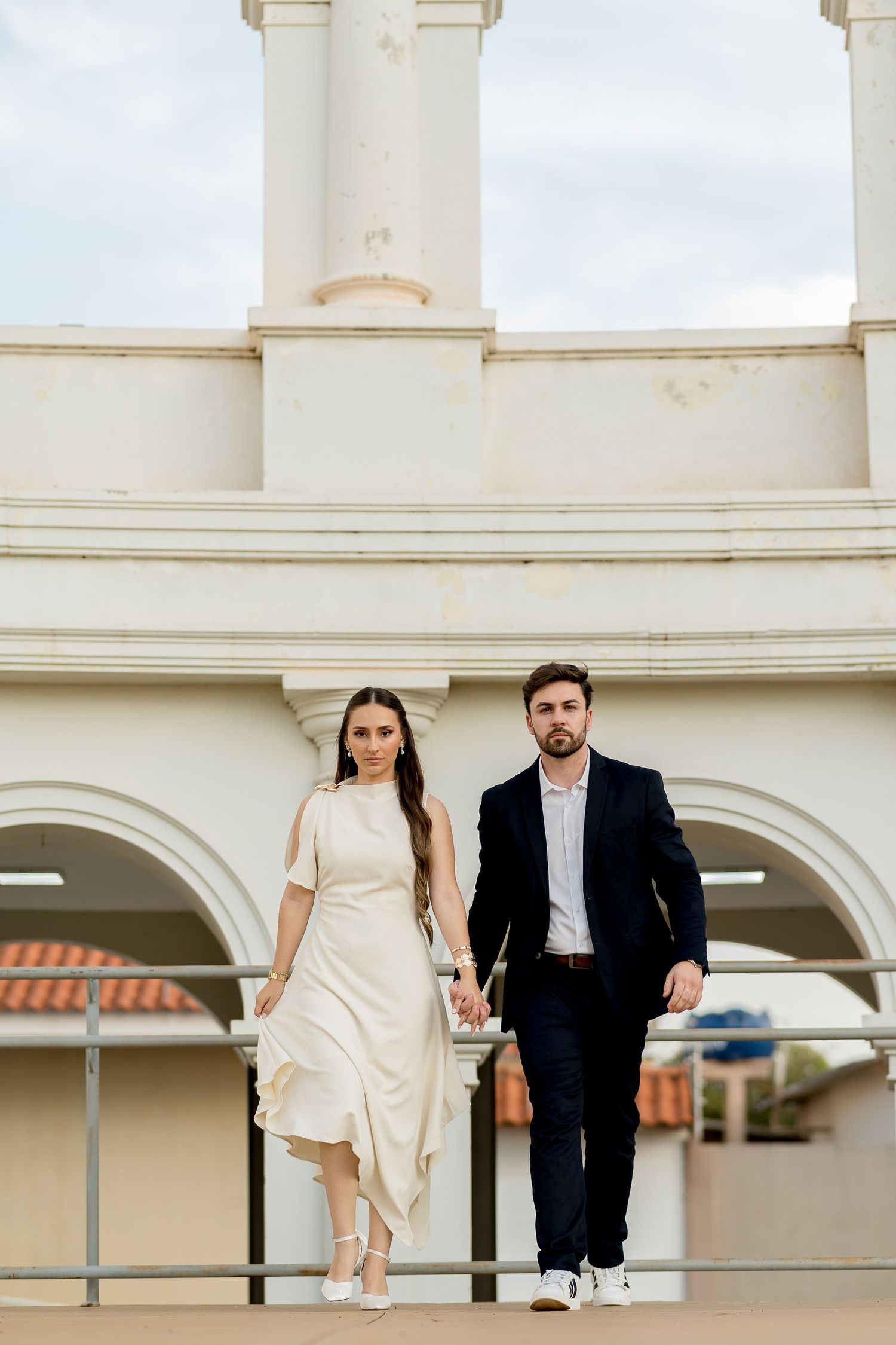 Elegant couple holding hands on a white architectural balcony, woman in white dress, man in navy suit.