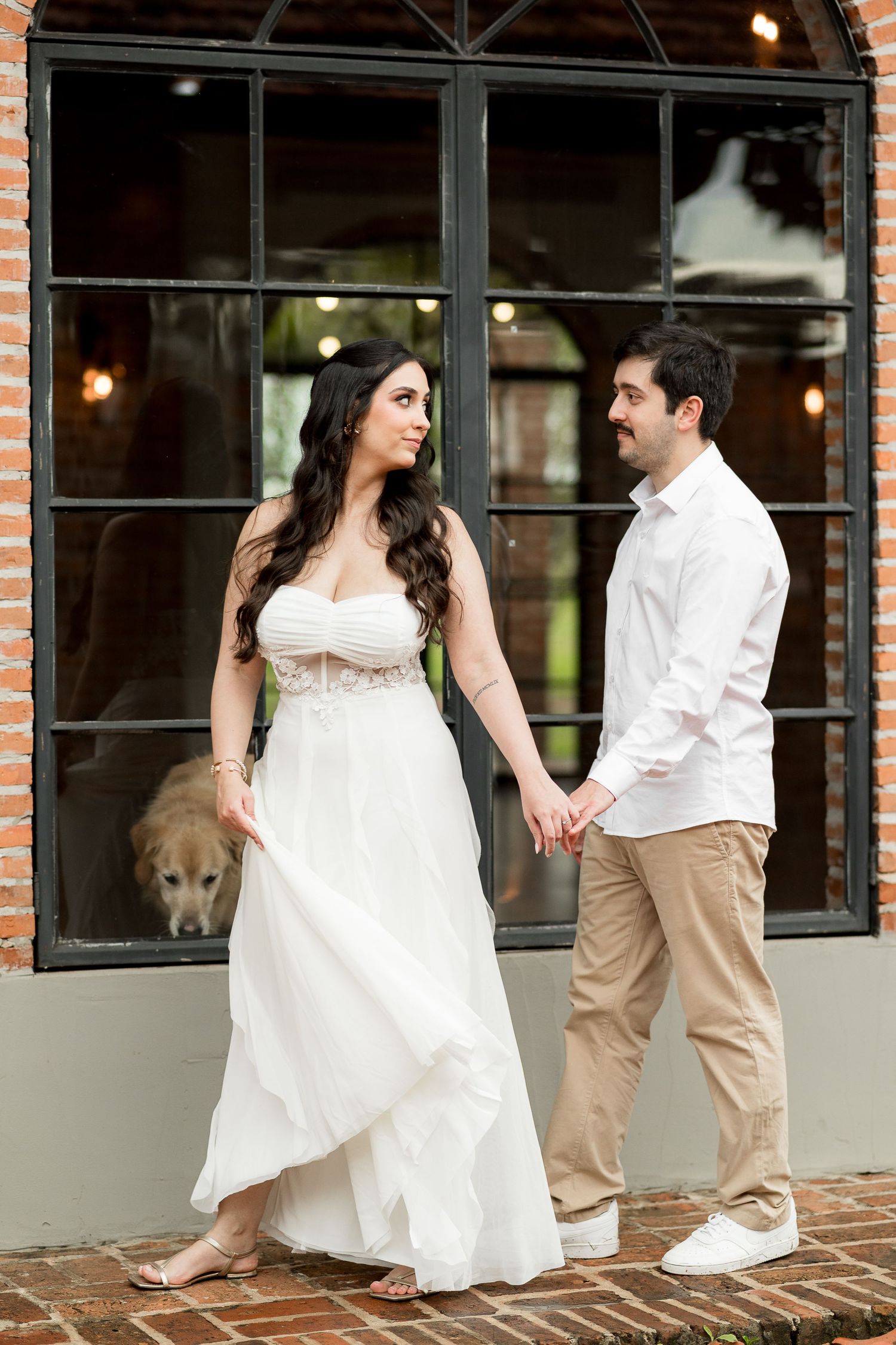 A couple in wedding attire holds hands outside a brick building, with a golden retriever visible through the window behind them.