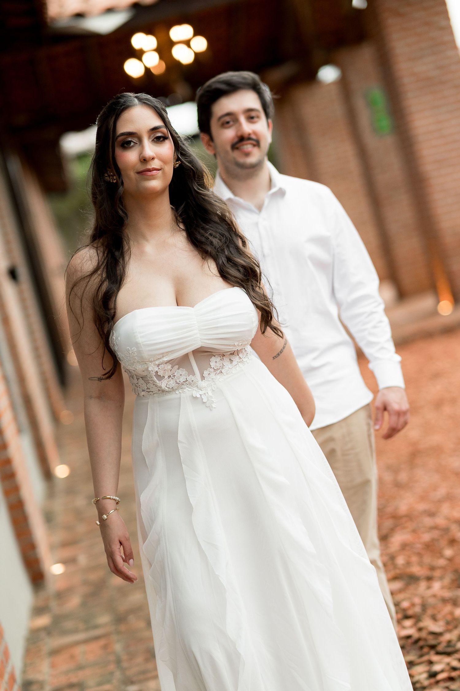 Elegant bride in strapless white gown walks with groom in white shirt at outdoor venue.