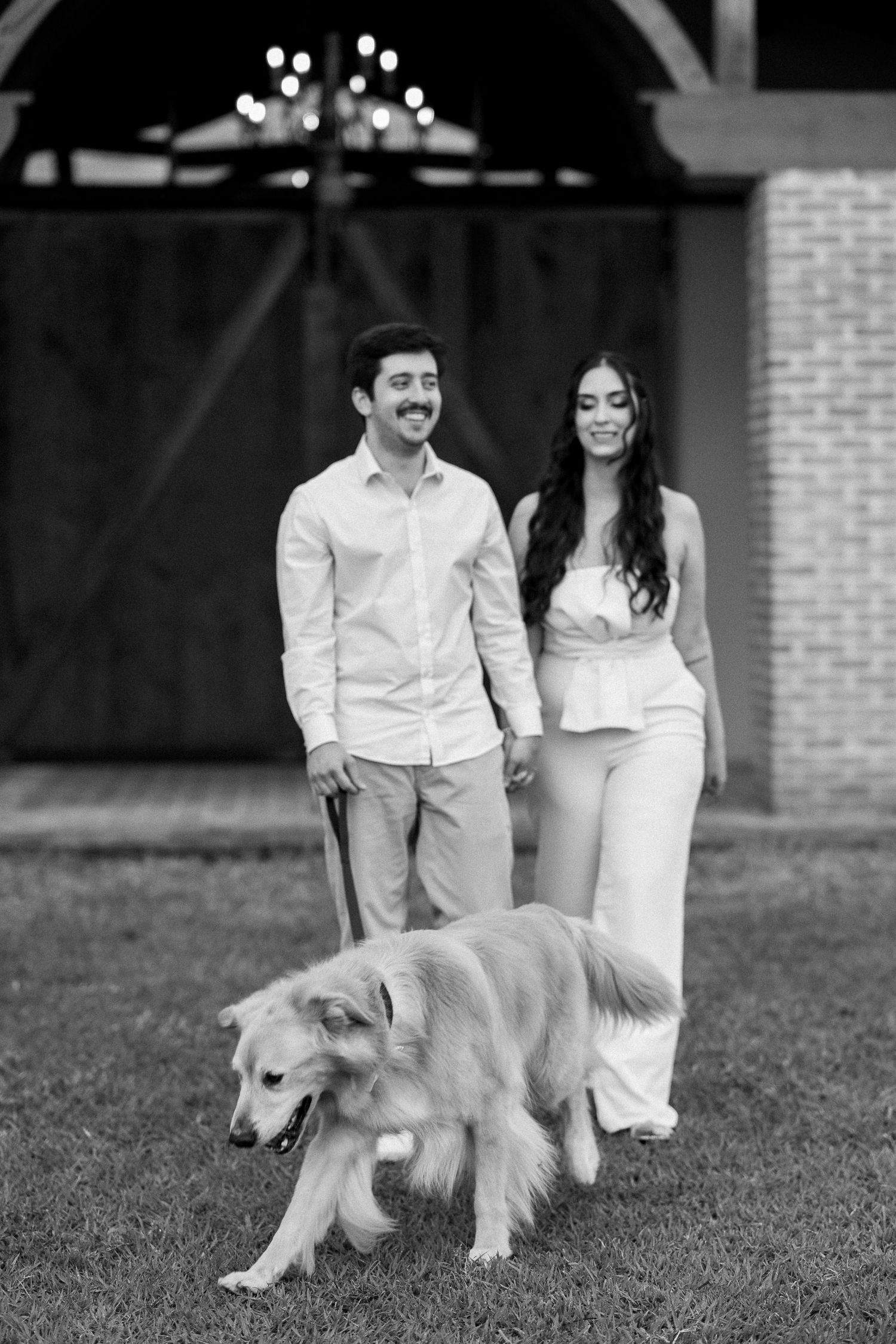 Black and white photo of a smiling couple walking their golden retriever on a leash outside a barn.