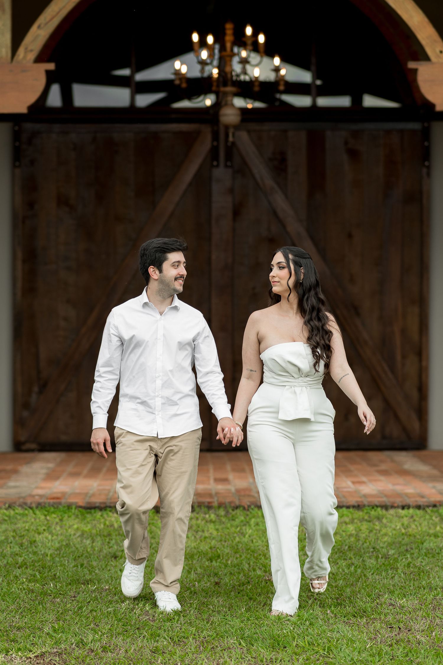 Couple holding hands walking in front of rustic barn doors, man in white shirt and woman in white strapless jumpsuit.