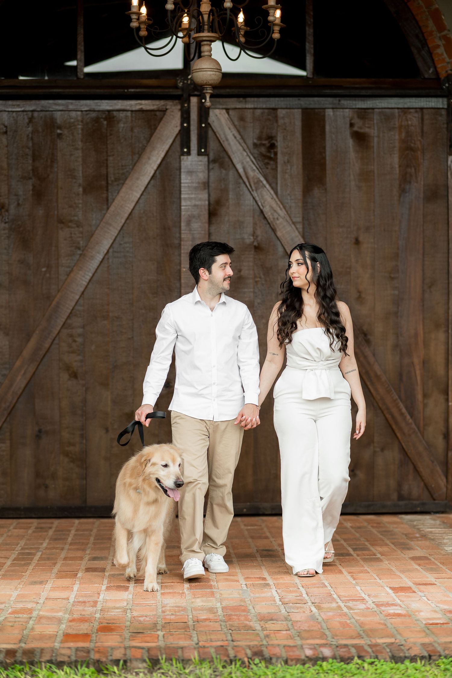 Couple holding hands walking golden retriever in front of rustic barn doors on brick pathway.