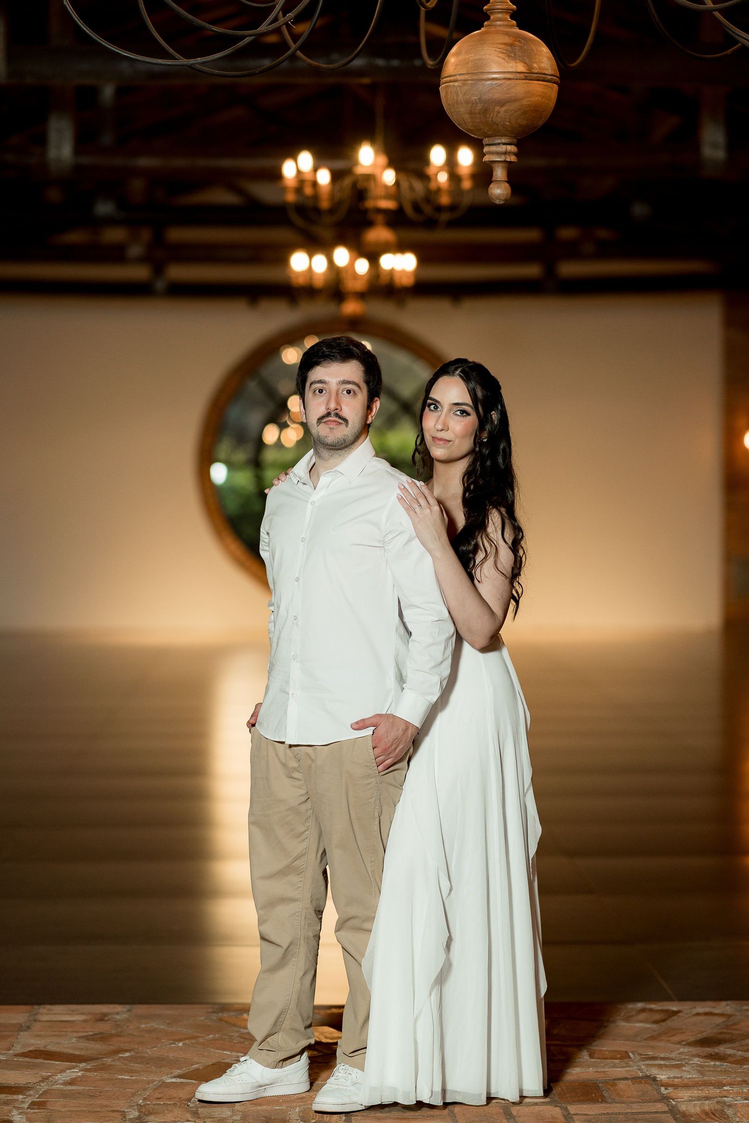 Elegant couple posing in a warmly lit venue, woman in white gown and man in white shirt with khaki pants.