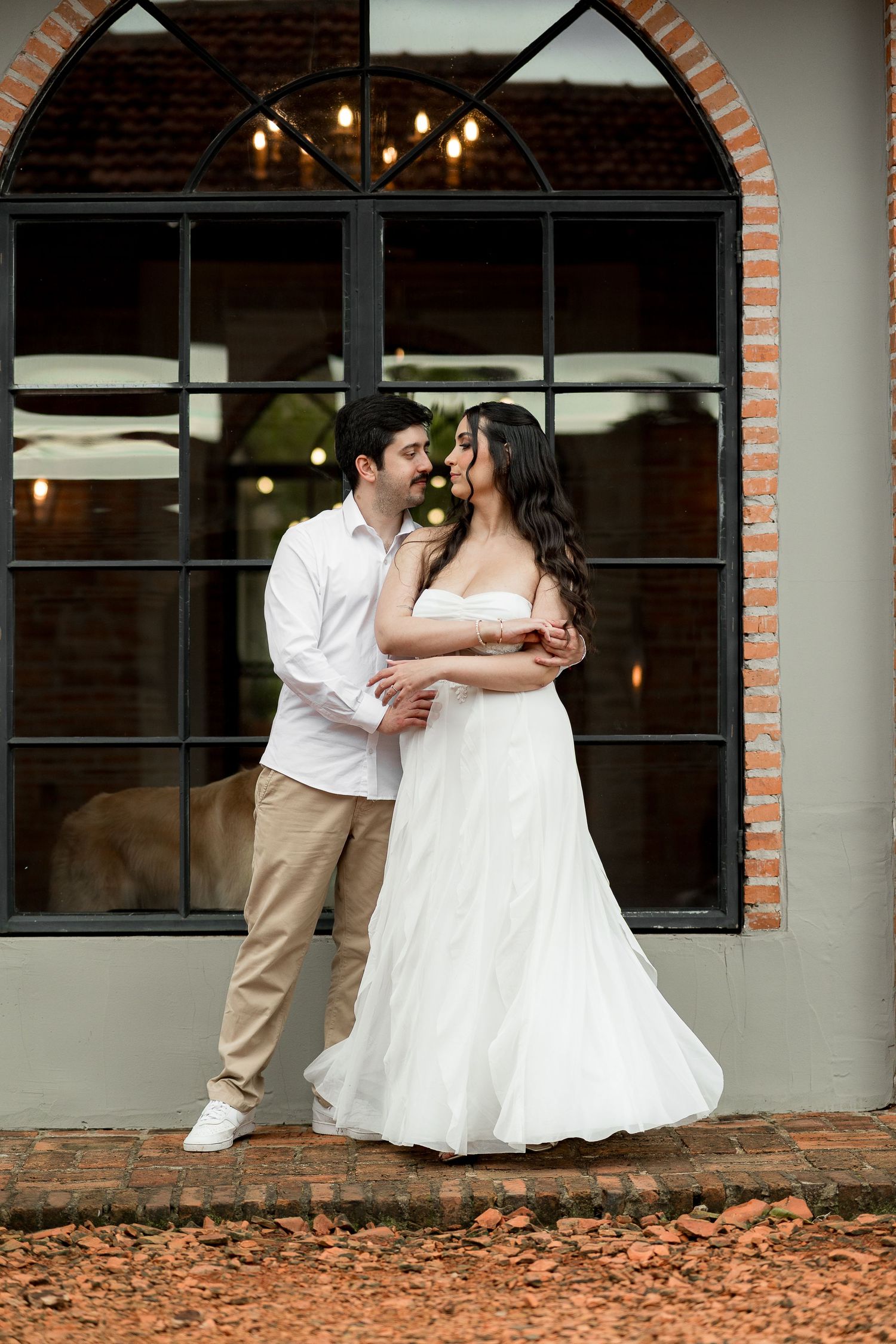 Couple in wedding attire embracing before arched brick window, bride in white gown, groom in white shirt and khakis.