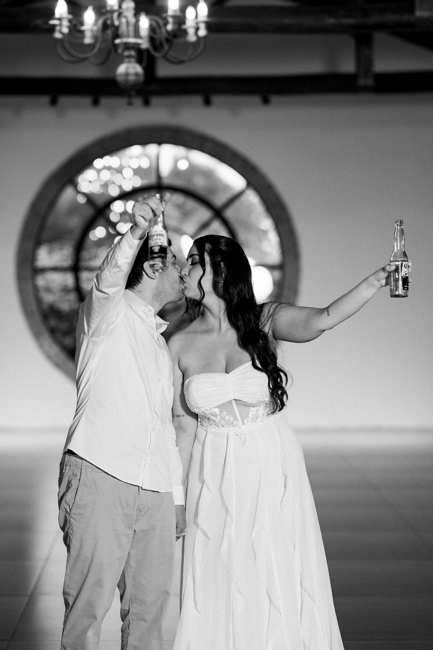 Black and white photo of a couple kissing and raising bottles at their wedding reception under a chandelier.