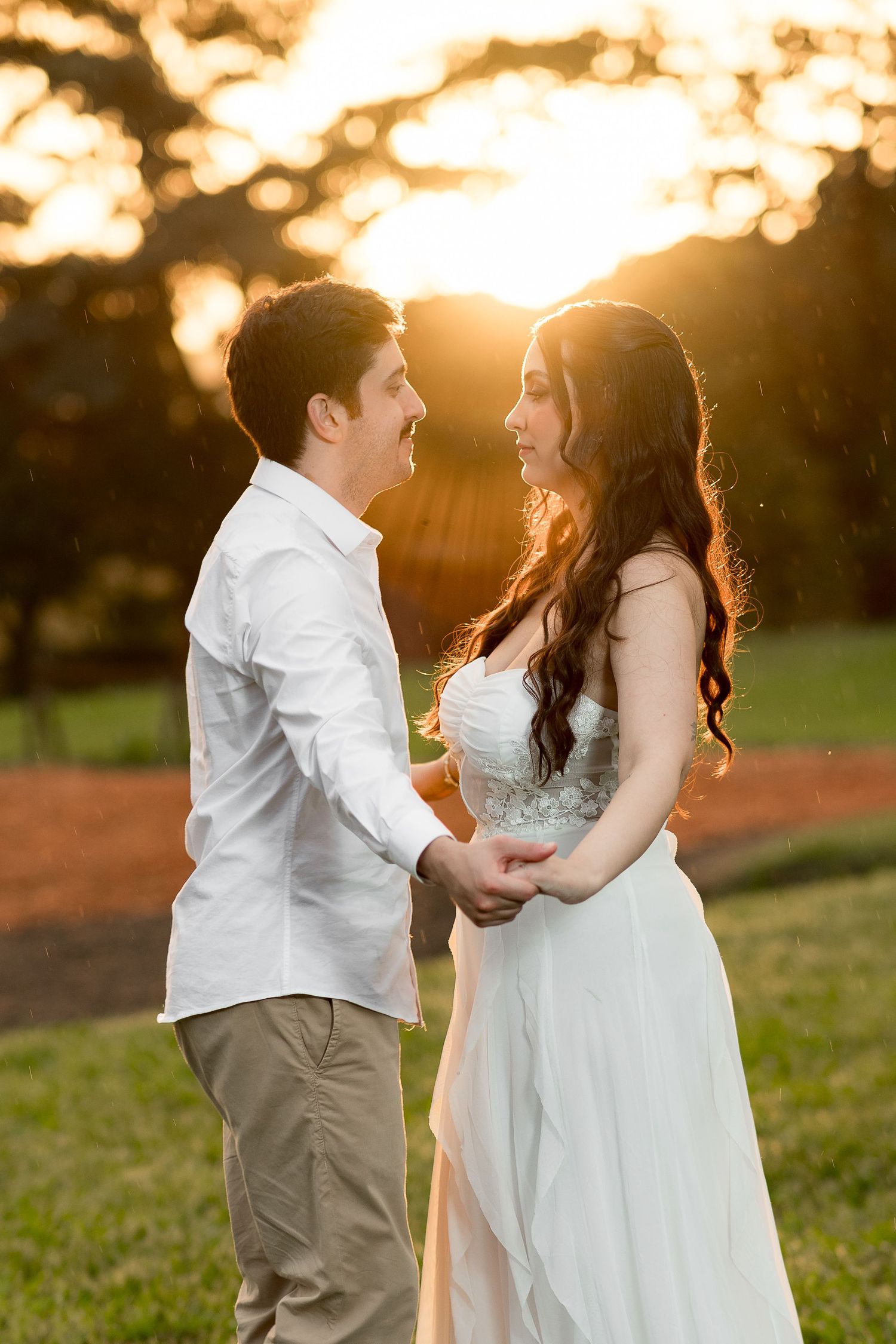 Couple holding hands at golden sunset outdoors, bride in white gown and groom in white shirt smiling at each other.