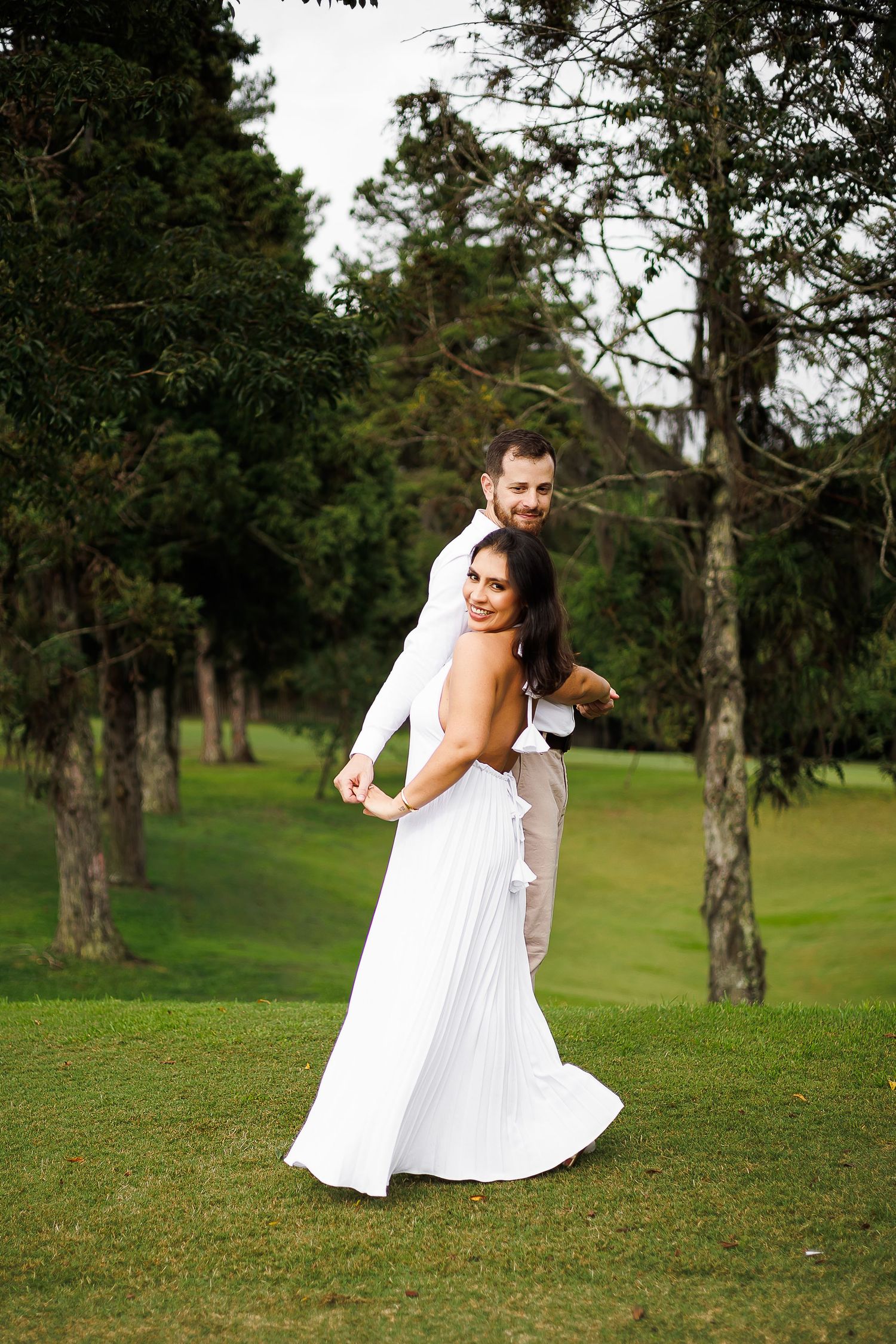 A smiling bride in a white gown embraced by her groom on a lush green lawn surrounded by tall trees.