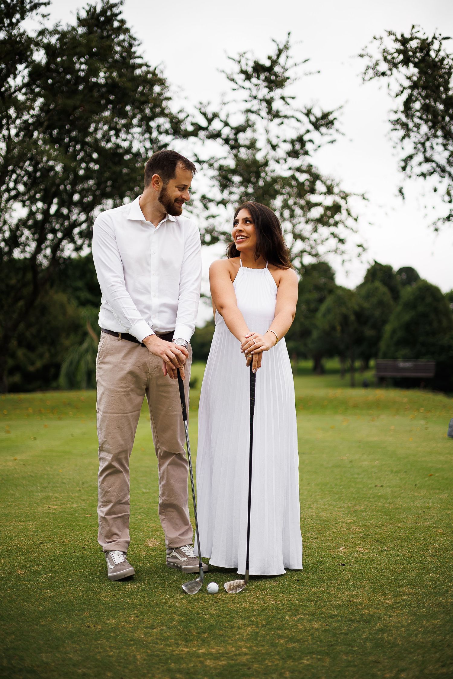 A couple in white attire smiling on a golf course, holding a golf club together on green grass.