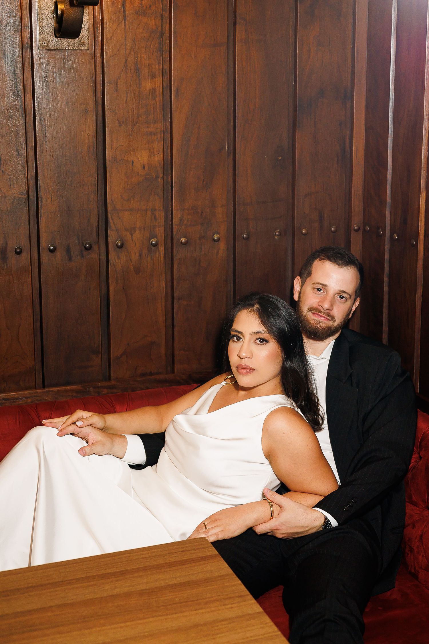 A bride in a white gown and groom in a black suit pose elegantly on a red velvet booth seat.