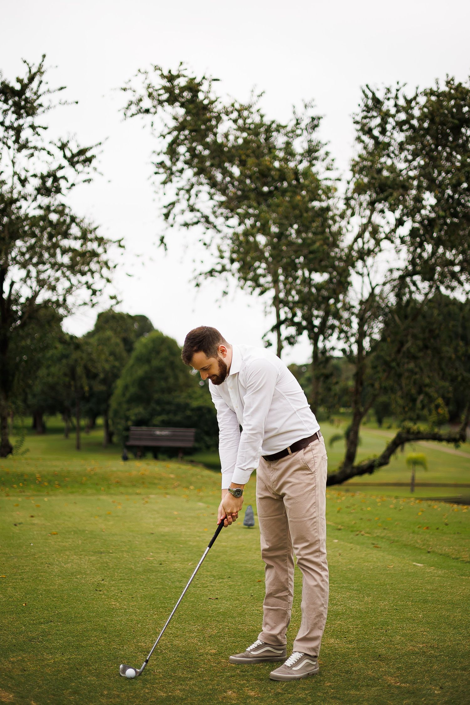 A bearded man in white shirt and khaki pants prepares to swing a golf club on a lush green golf course.