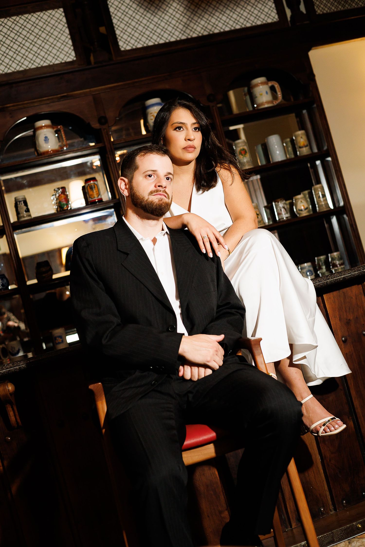Couple posing elegantly, man in black suit seated, woman in white outfit resting arm on his shoulder in a warmly lit room.