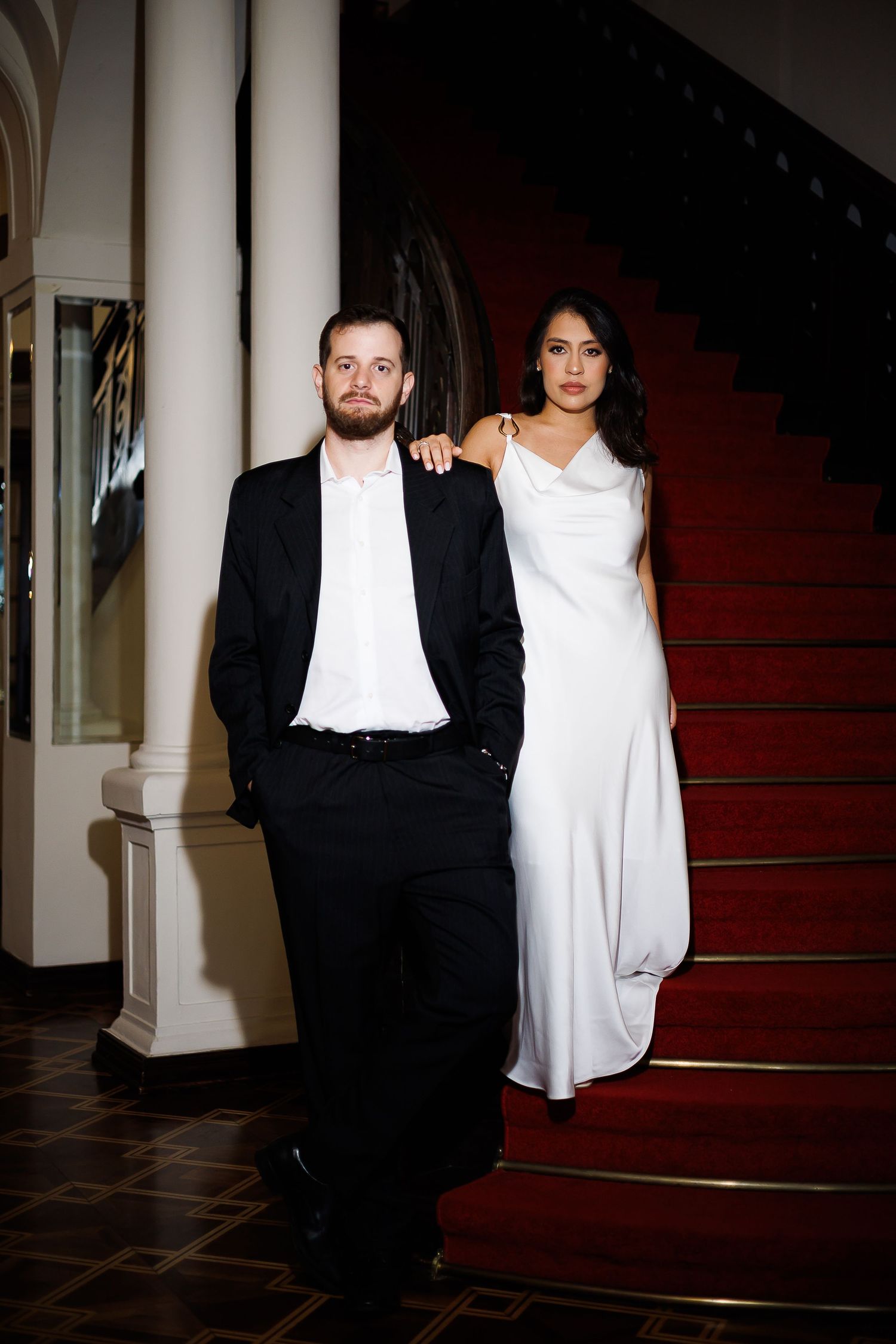 Elegant couple posing on red carpeted staircase, man in black suit, woman in white satin gown.
