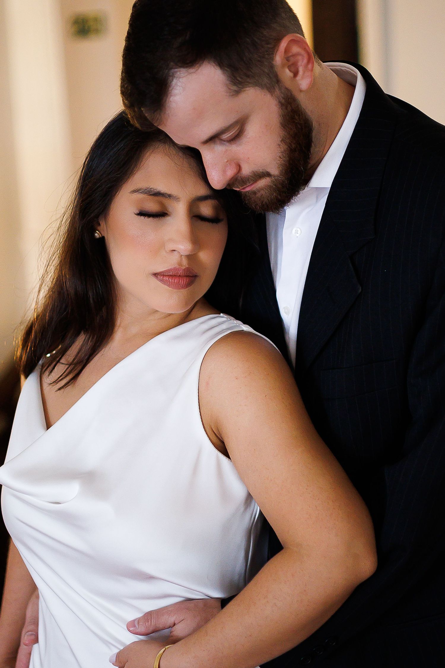 A couple in formal wedding attire embraces tenderly, the bride in white satin and groom in black suit, eyes closed.