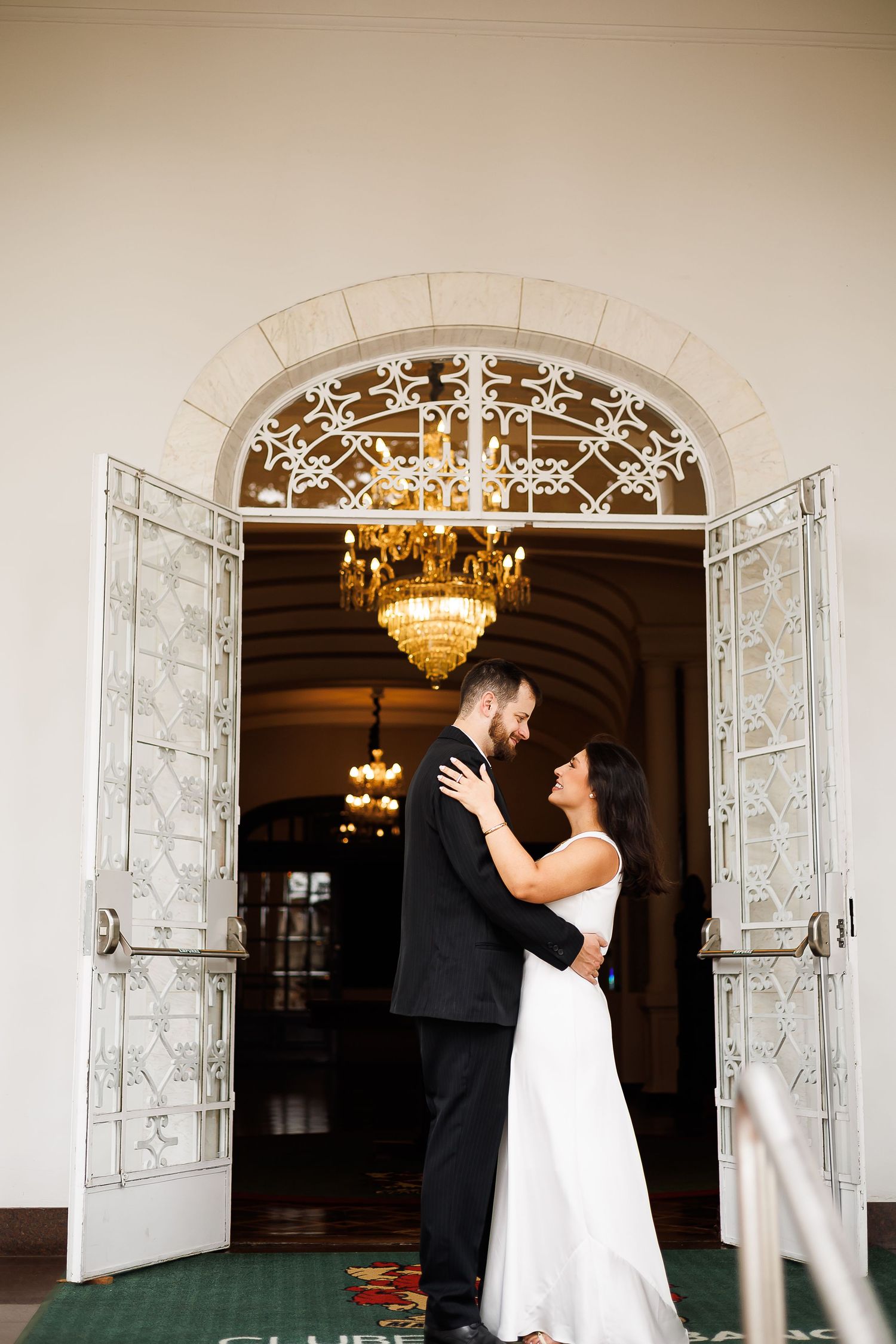 Bride and groom embrace in elegant arched doorway with ornate ironwork and chandelier.