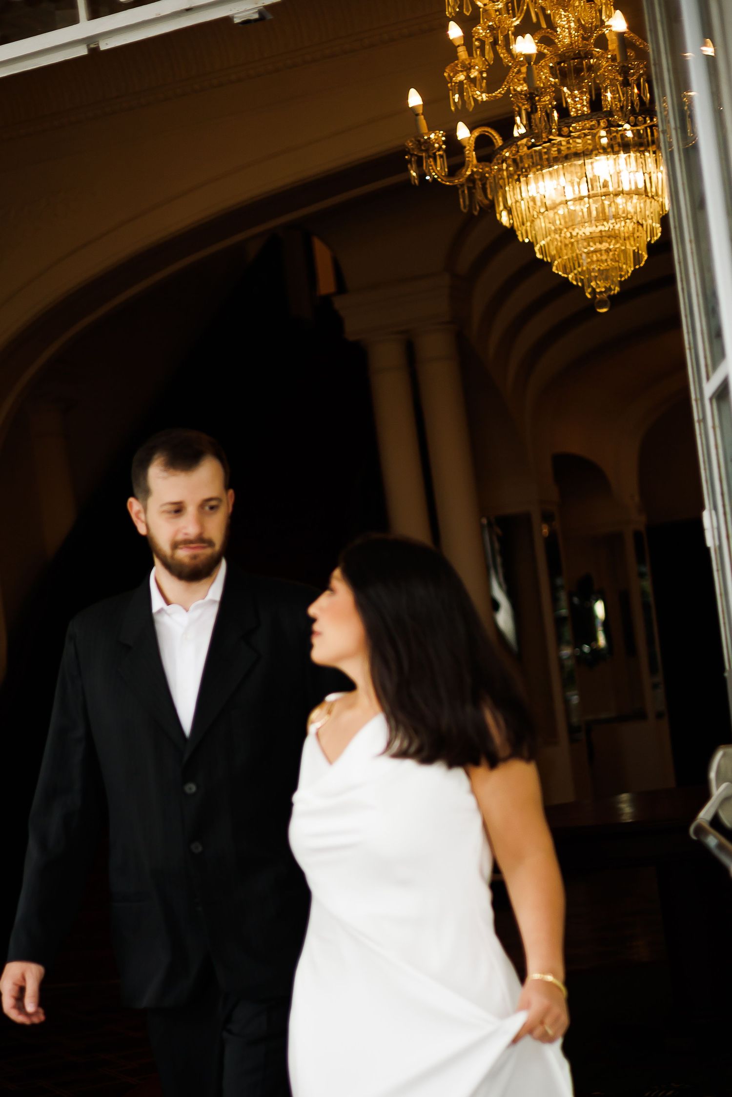 Elegant couple in formal attire walking under a grand chandelier in a dimly lit ballroom.