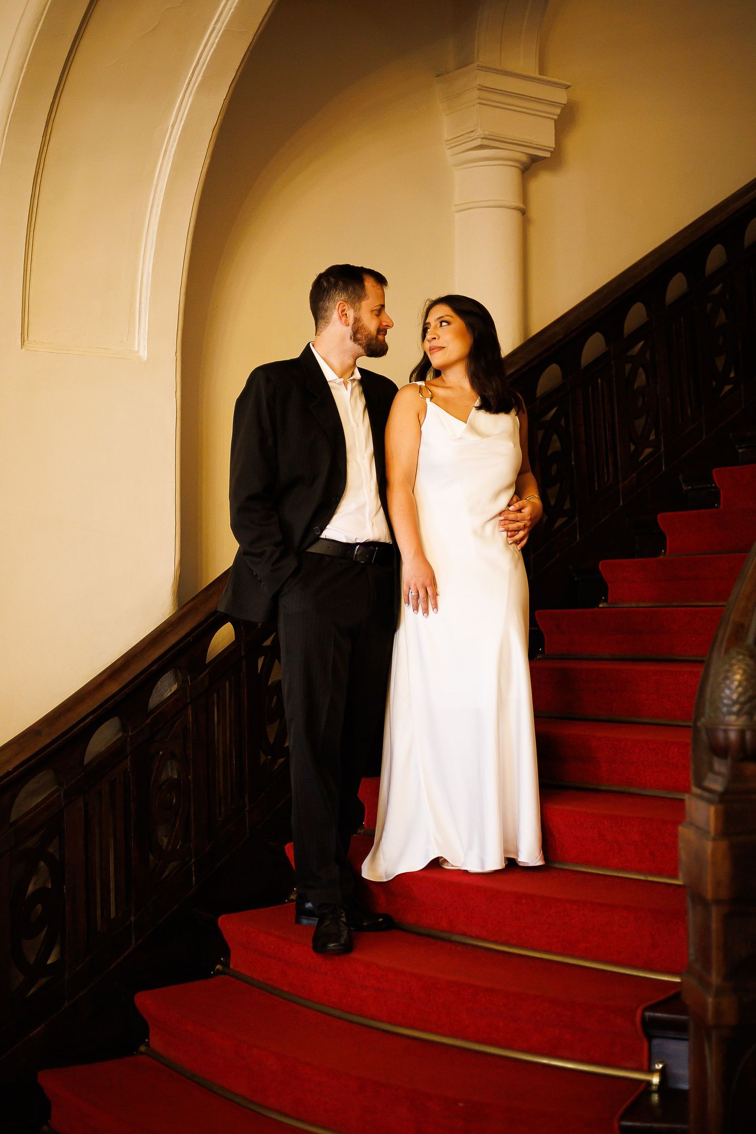 Couple in wedding attire posing on red-carpeted grand staircase, bride in white gown and groom in black suit.