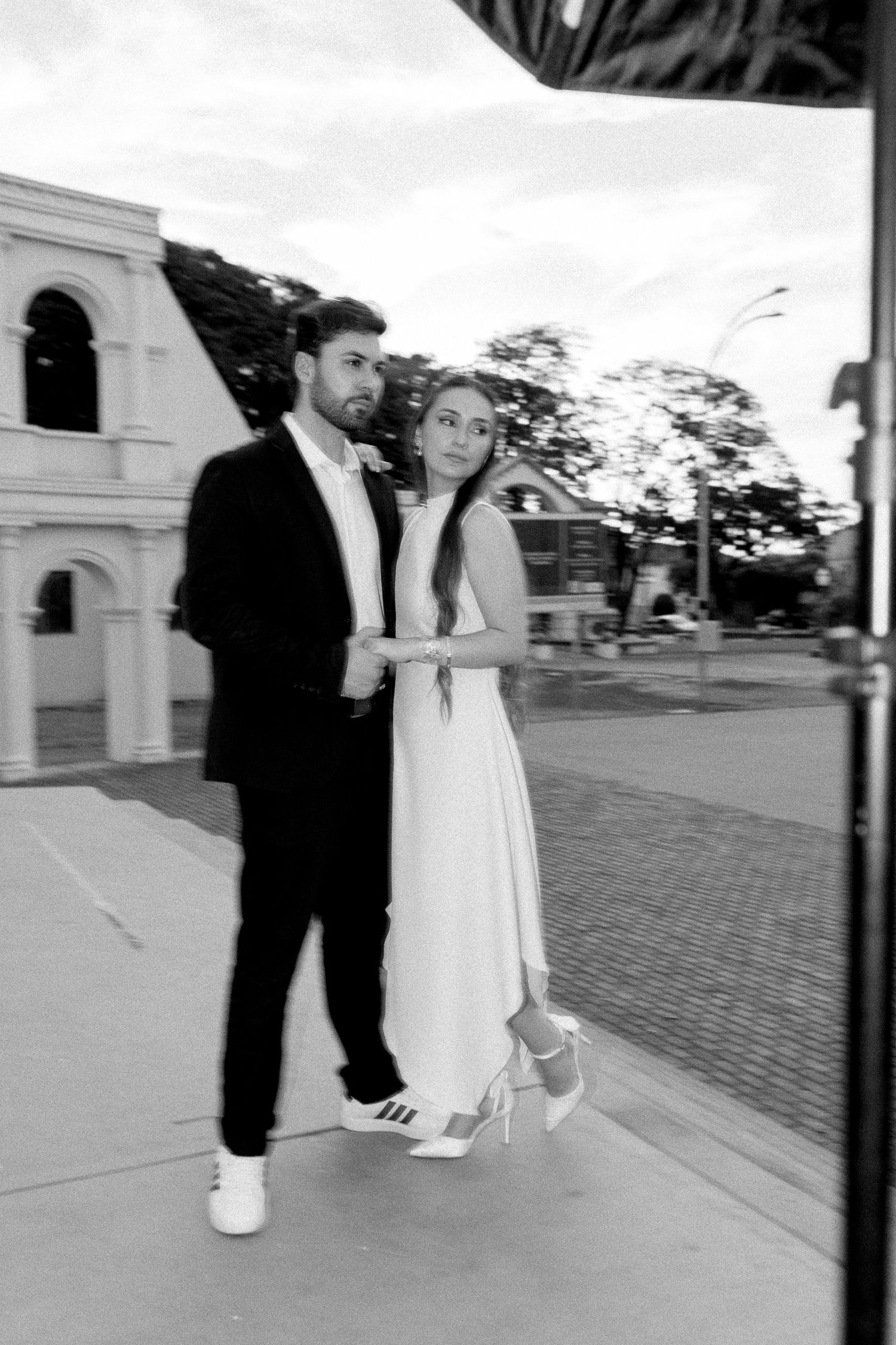 Black and white photo of a couple in wedding attire posing on a sidewalk, bride in white dress with Adidas sneakers.