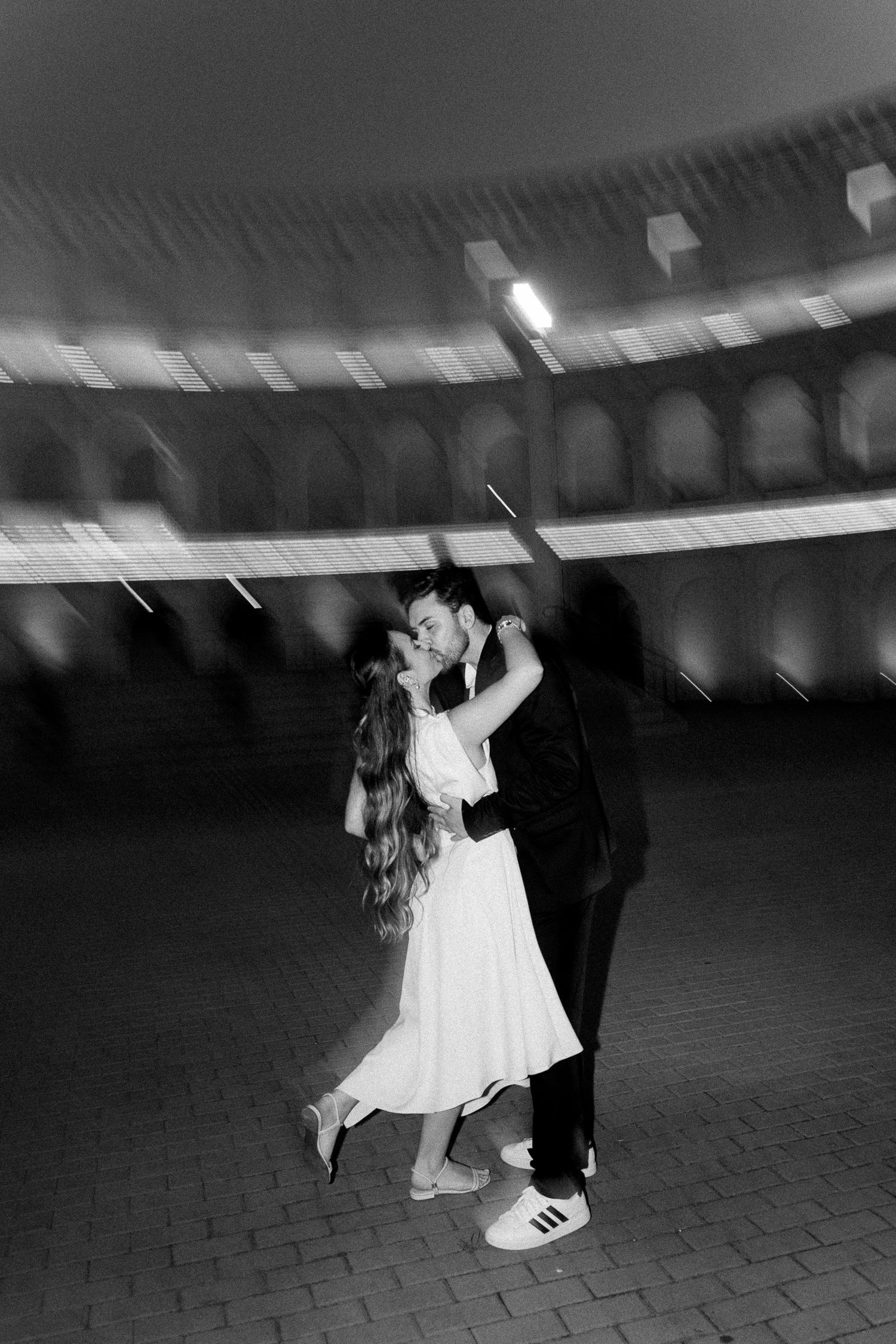 Black and white photo of a couple kissing and dancing outdoors at night near an illuminated arched building.