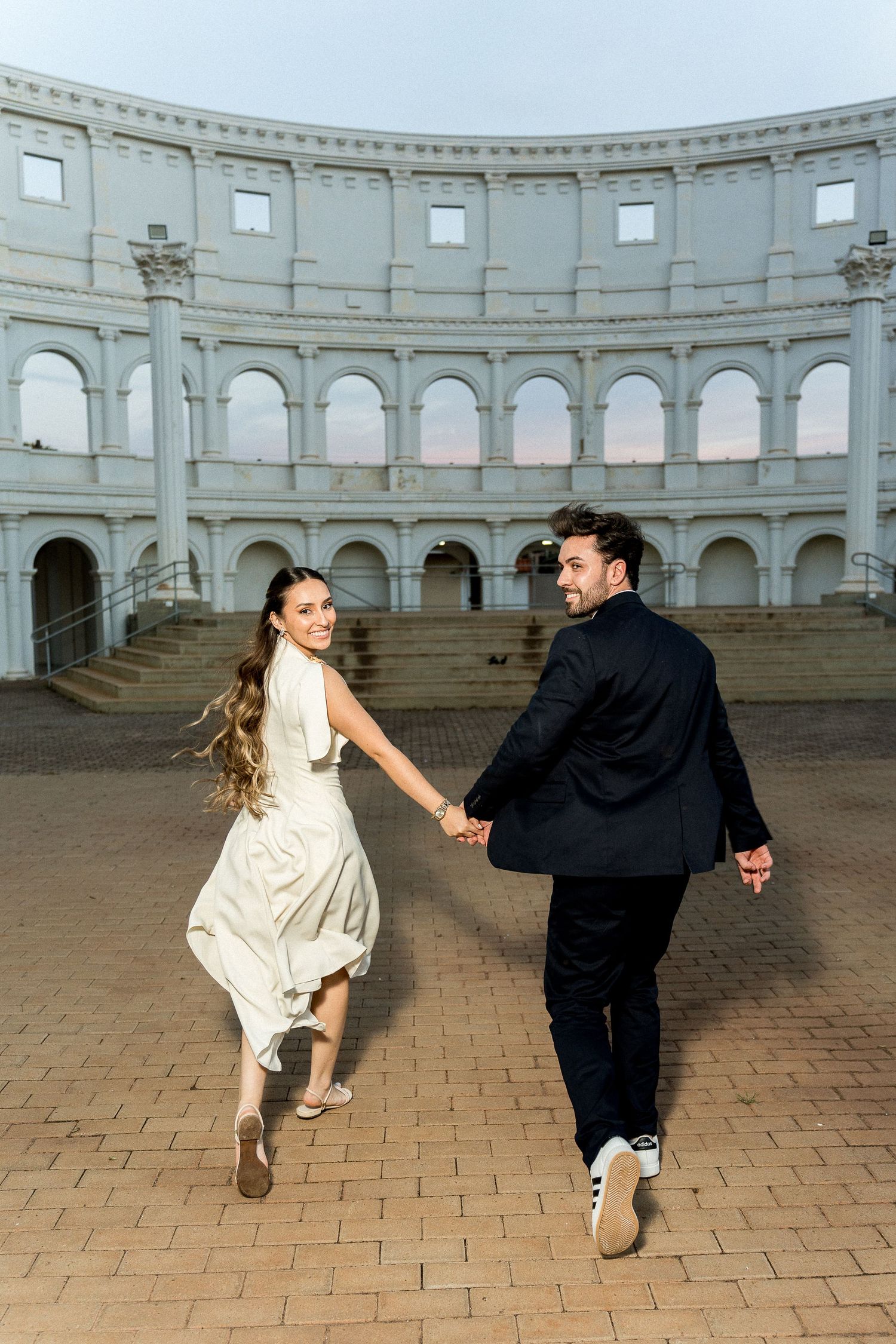 Couple in wedding attire holding hands and running in front of a grand Roman-style amphitheater at dusk.