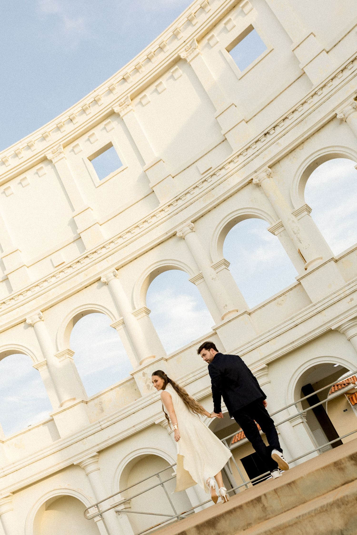 Couple in wedding attire posing on steps in front of the Roman Colosseum on a sunny day.