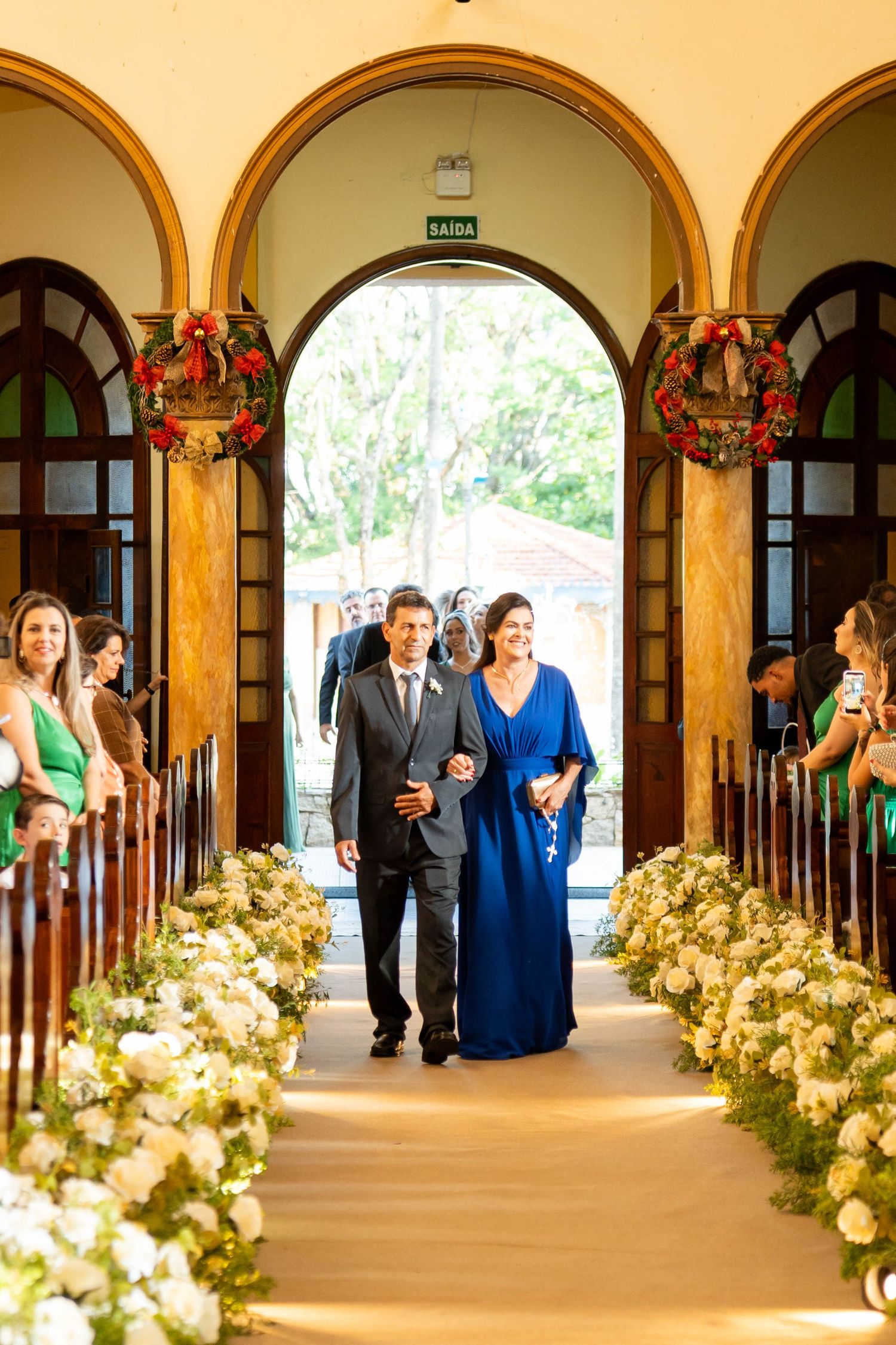 Couple walks down church aisle lined with white floral arrangements, woman in blue gown, Christmas wreaths on doors.