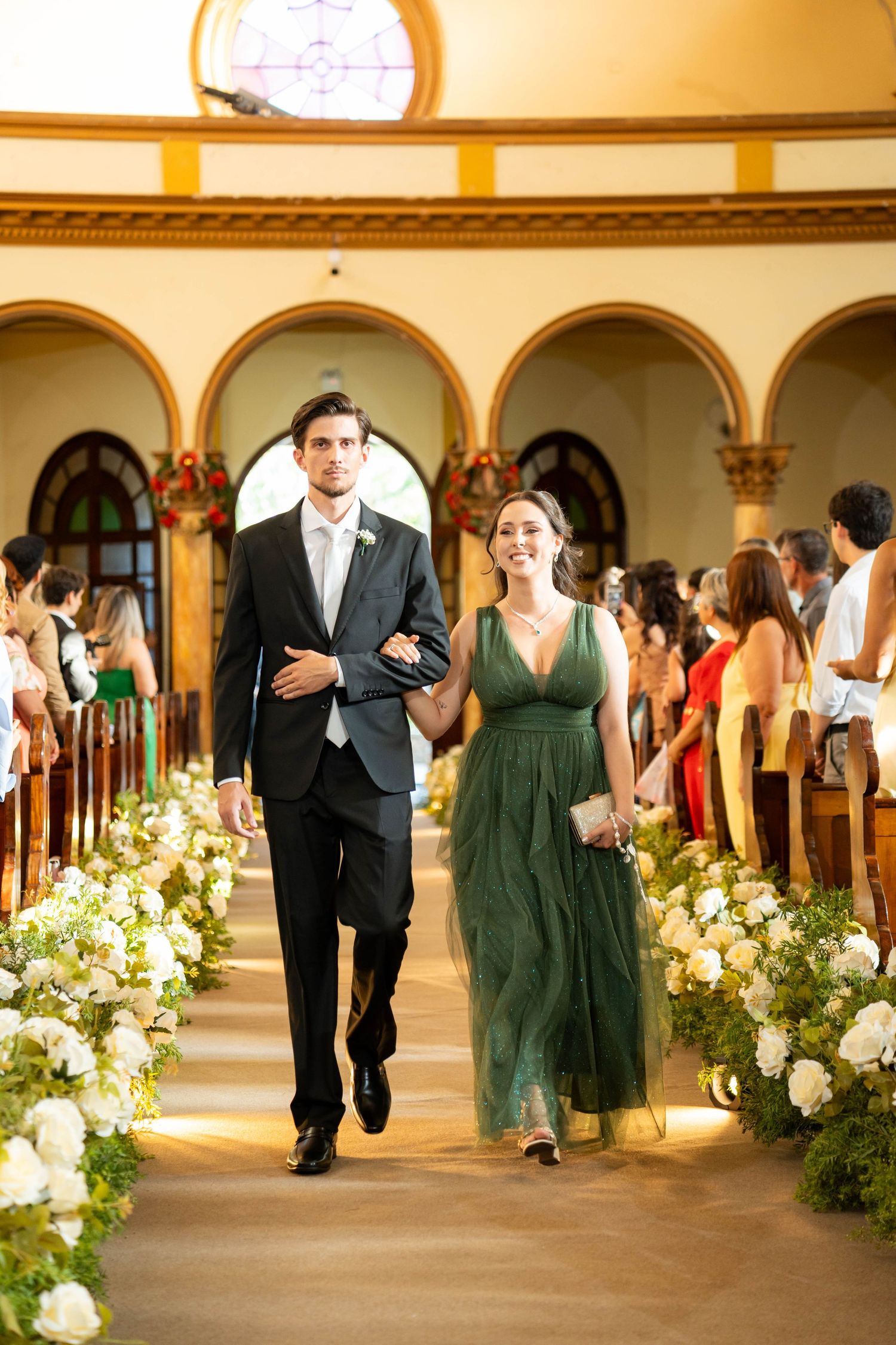 A couple walks down a flower-lined church aisle, man in black suit and woman in elegant green gown, during a wedding ceremony.