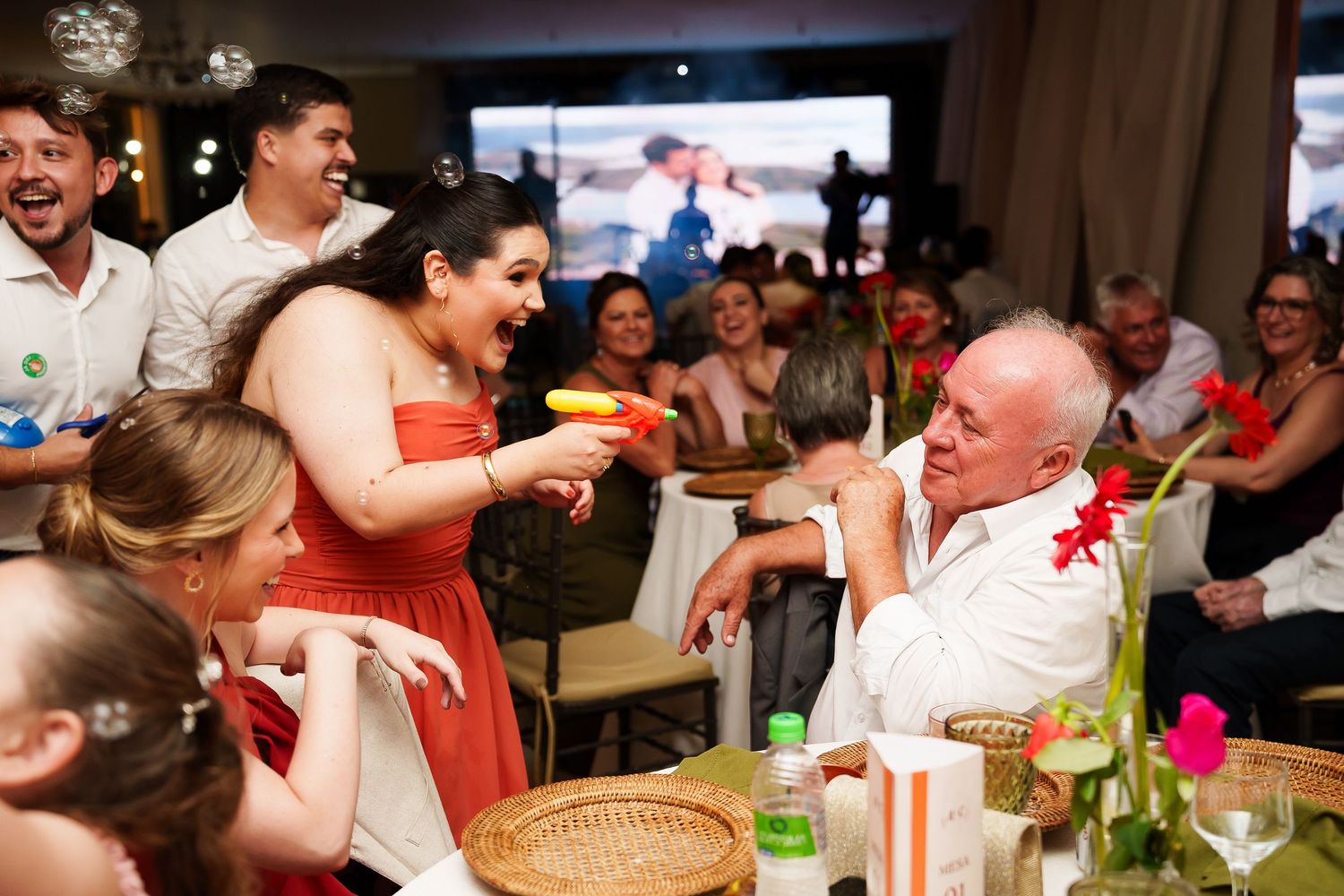 A woman in a red dress playfully interacts with an older man at a lively wedding reception dinner party.