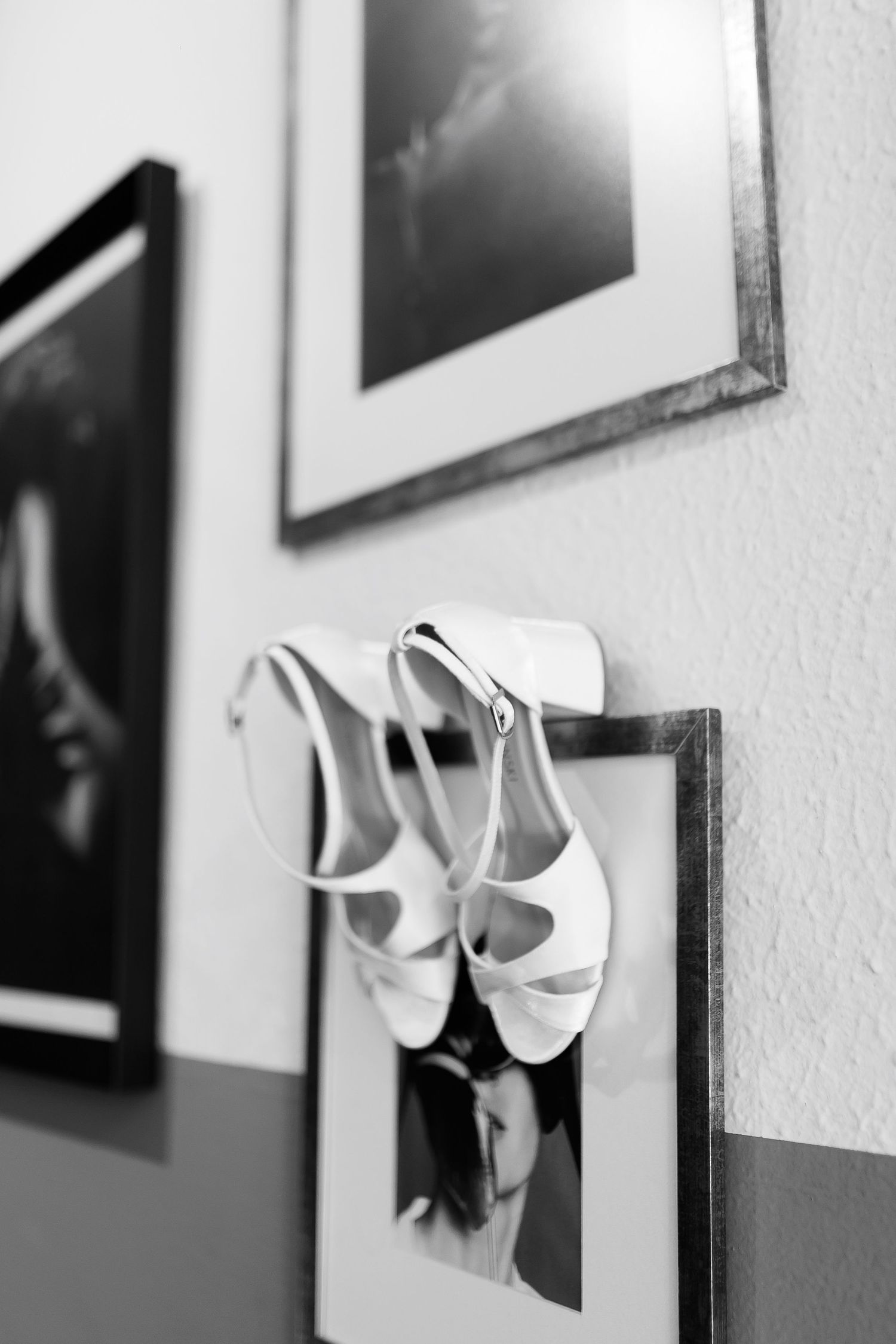 Black and white photo of white bridal heels hanging on a picture frame on a wall with art prints.
