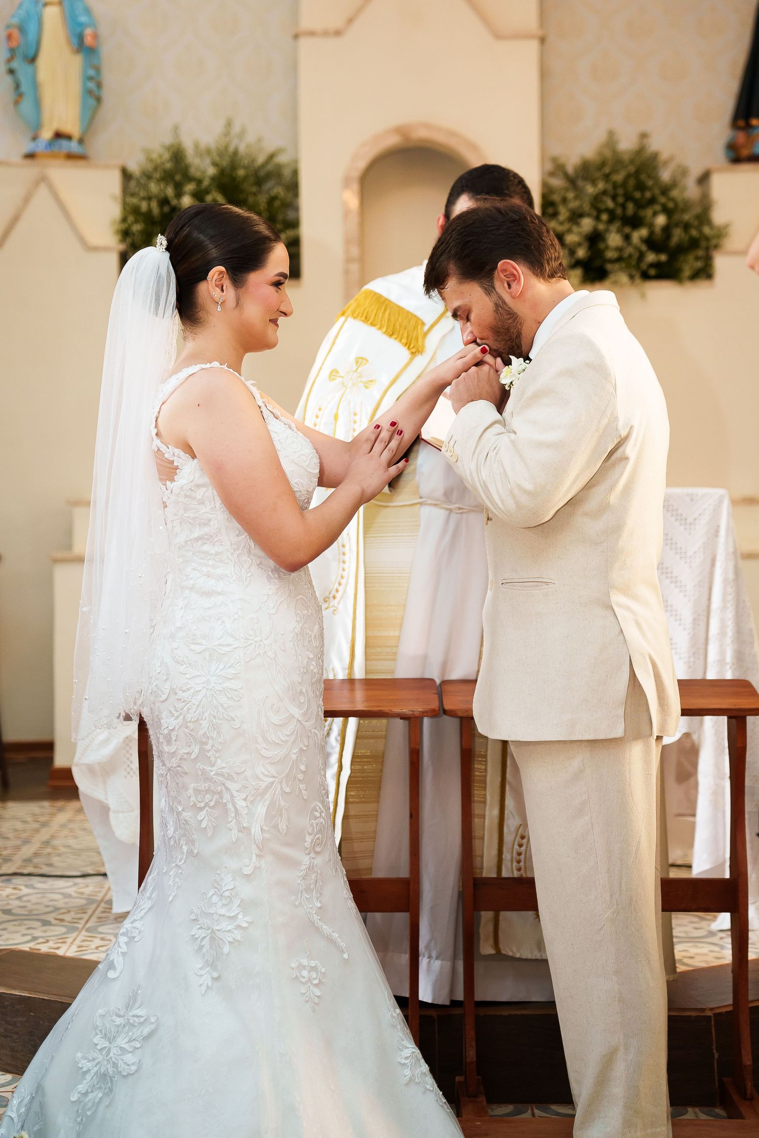 Groom kissing bride's hand at church altar during wedding ceremony, both dressed in white.