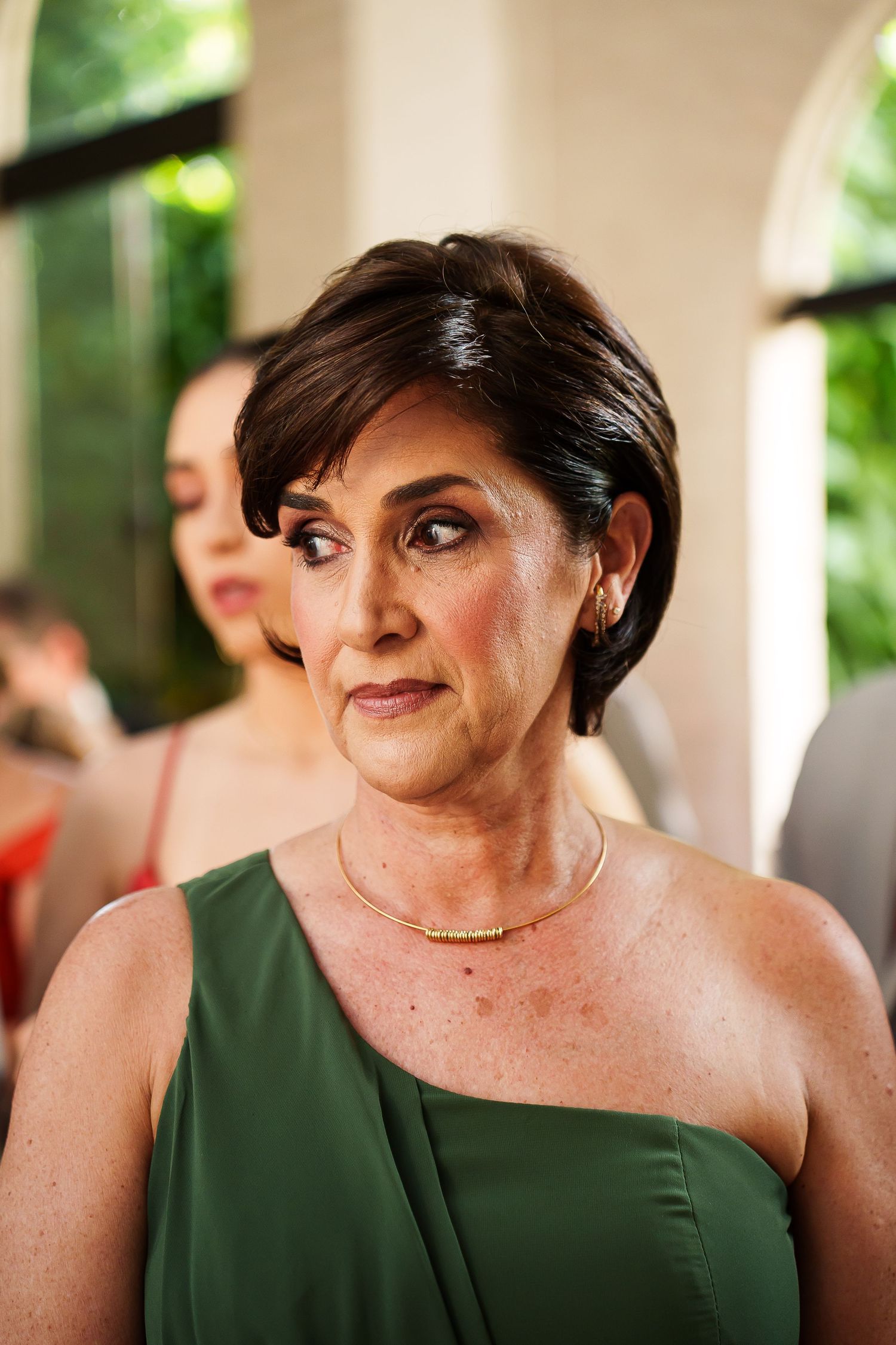 Elegant older woman in green one-shoulder dress with gold necklace at formal event.