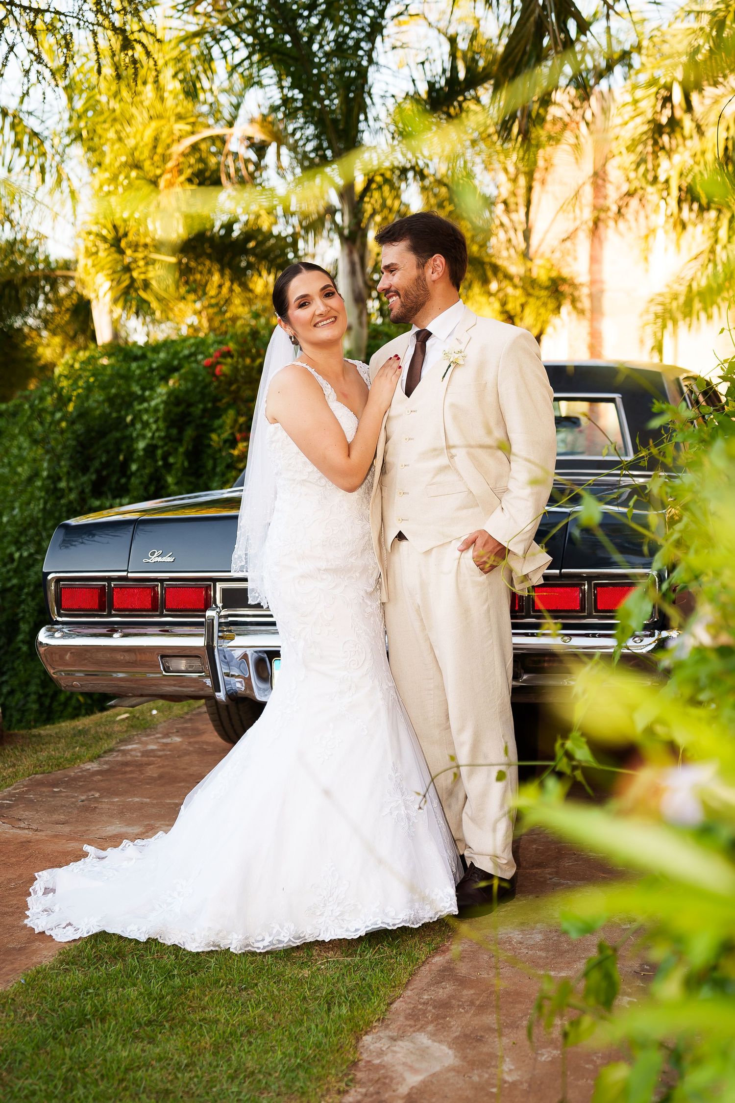 Bride in white lace gown and groom in cream suit pose by vintage black car surrounded by lush greenery.