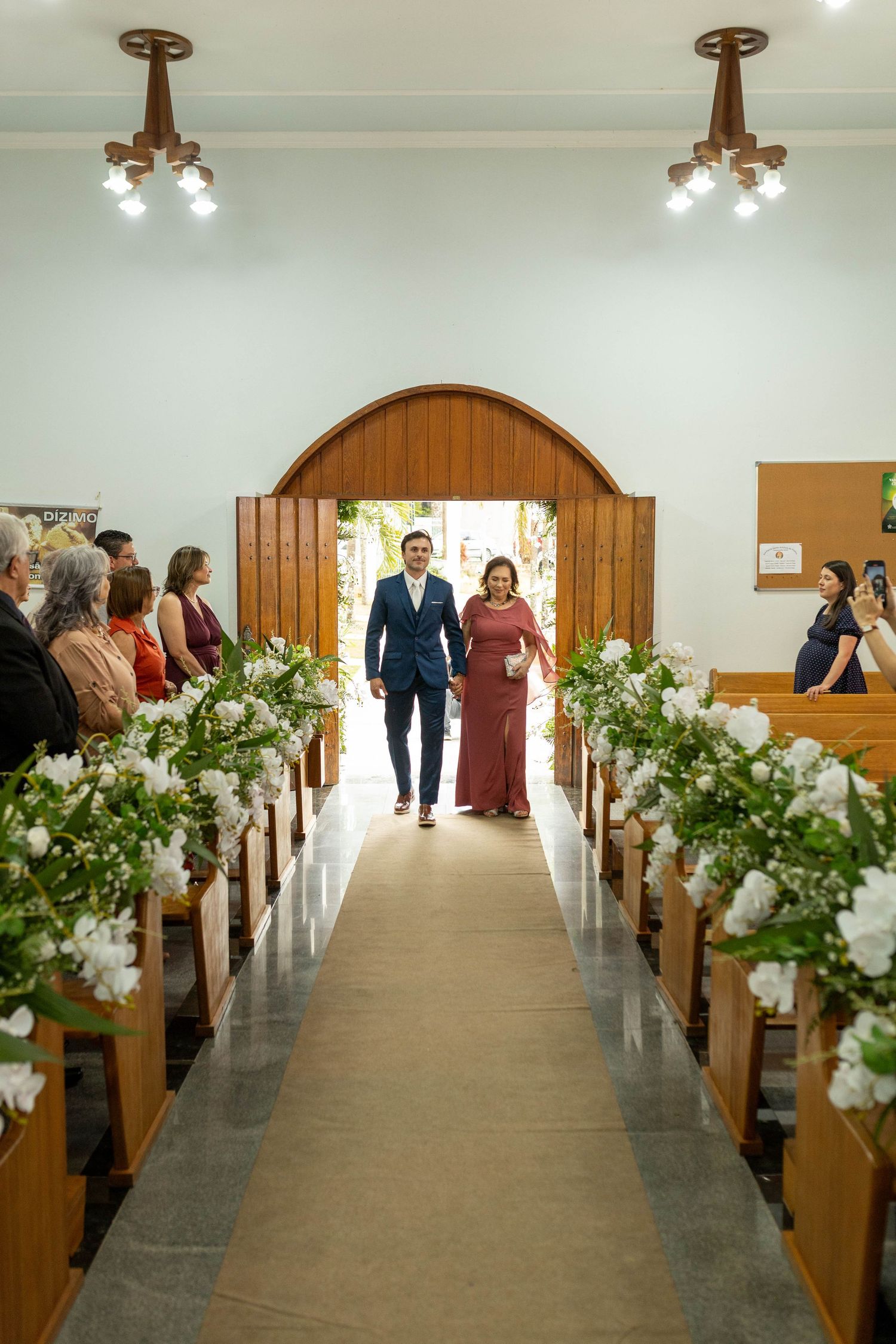 Groom in blue suit walks down church aisle with woman in burgundy dress, flanked by white floral arrangements on wooden pews.