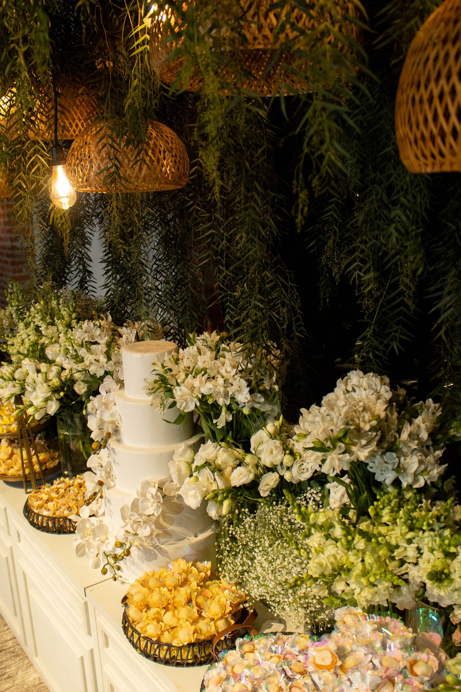 Elegant wedding dessert table with white tiered cake, white floral arrangements, and assorted sweets under warm pendant lights.
