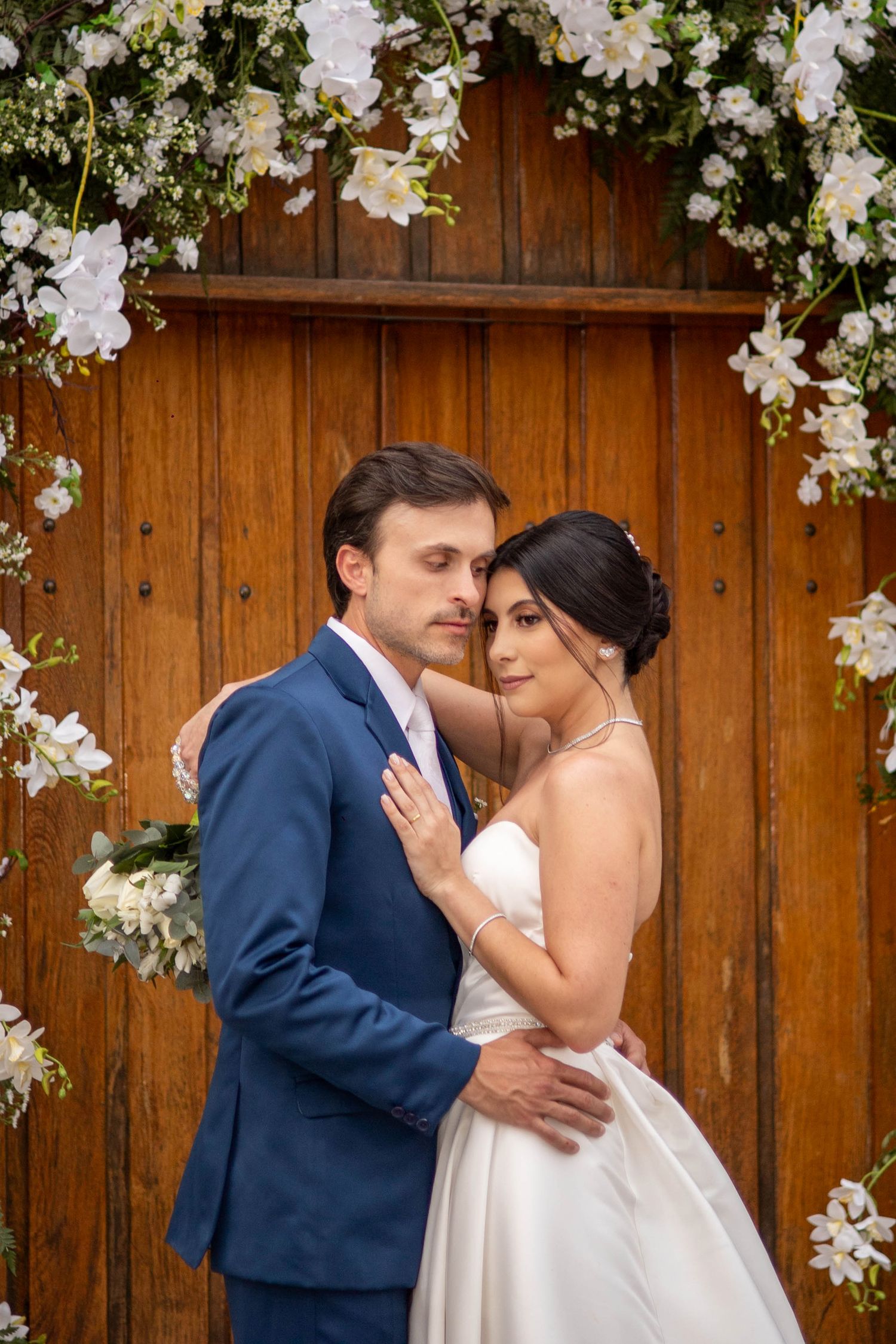 Bride and groom embracing under a floral arch with white flowers against a rustic wooden door backdrop.