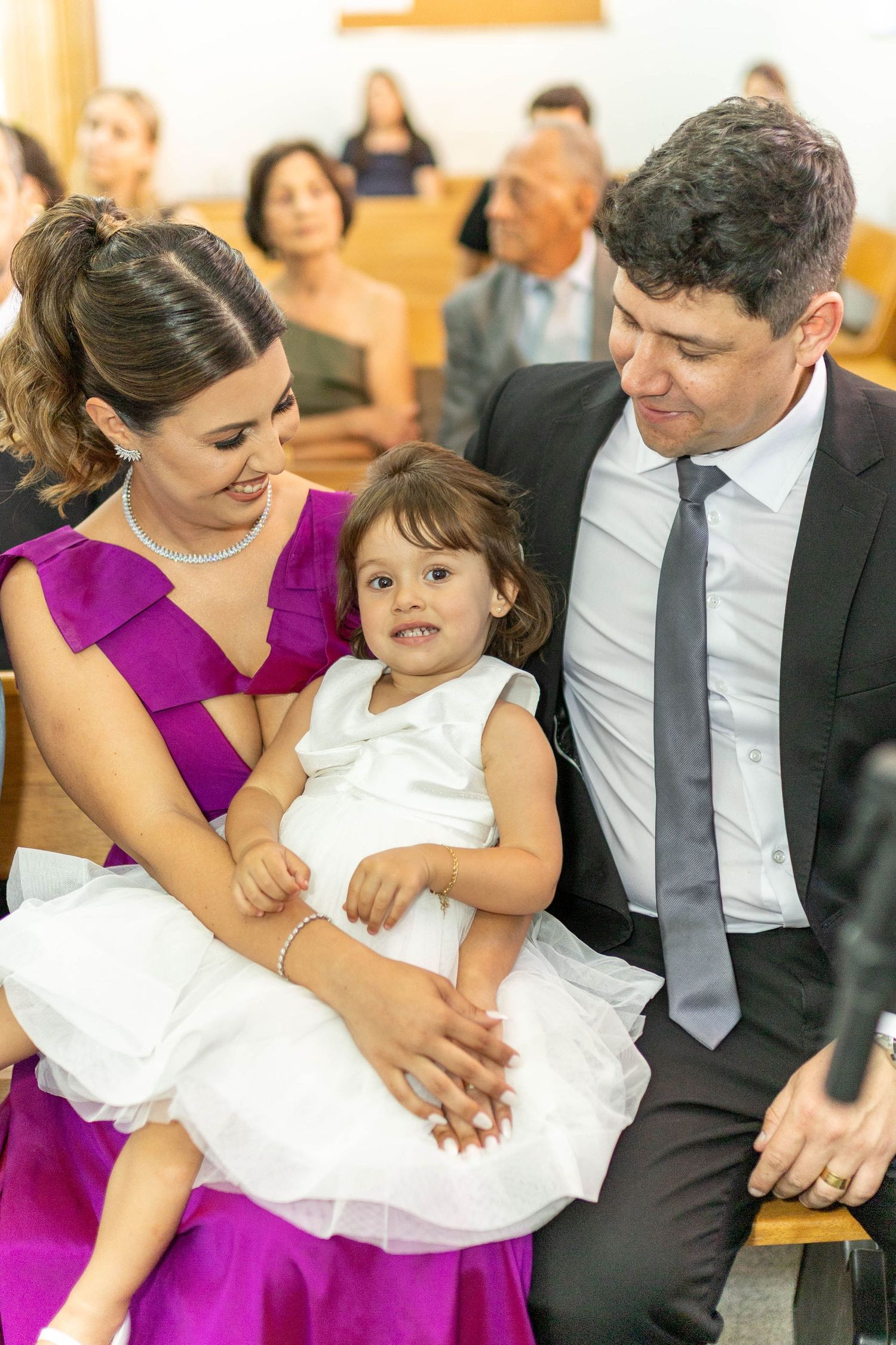 Family in church, woman in purple dress holds toddler in white dress beside man in suit.