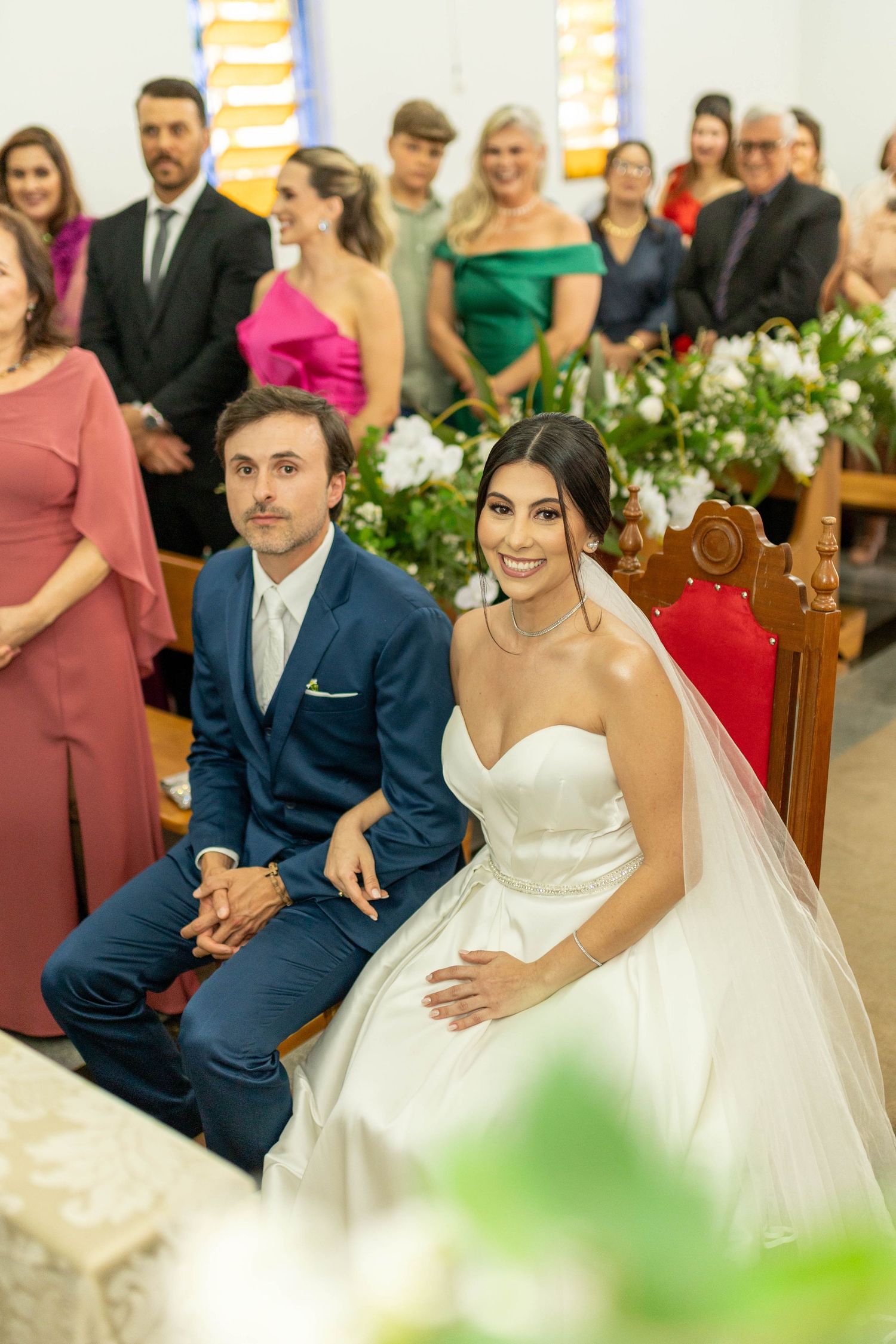Bride in white gown and groom in blue suit seated at church altar during wedding ceremony with guests in background.